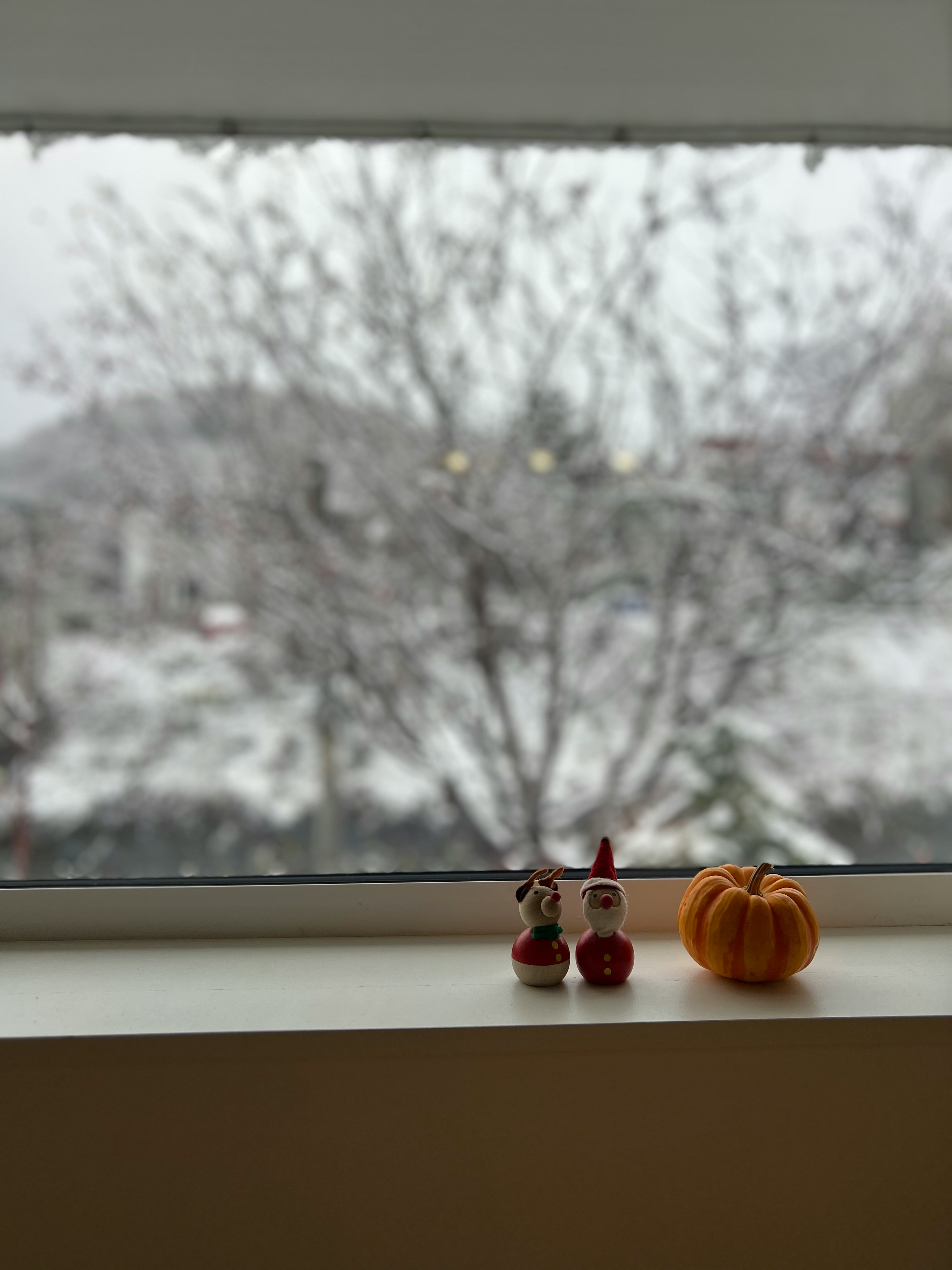 Foreground features red Santa figurines and an orange pumpkin on a windowsill with a snowy landscape in the background