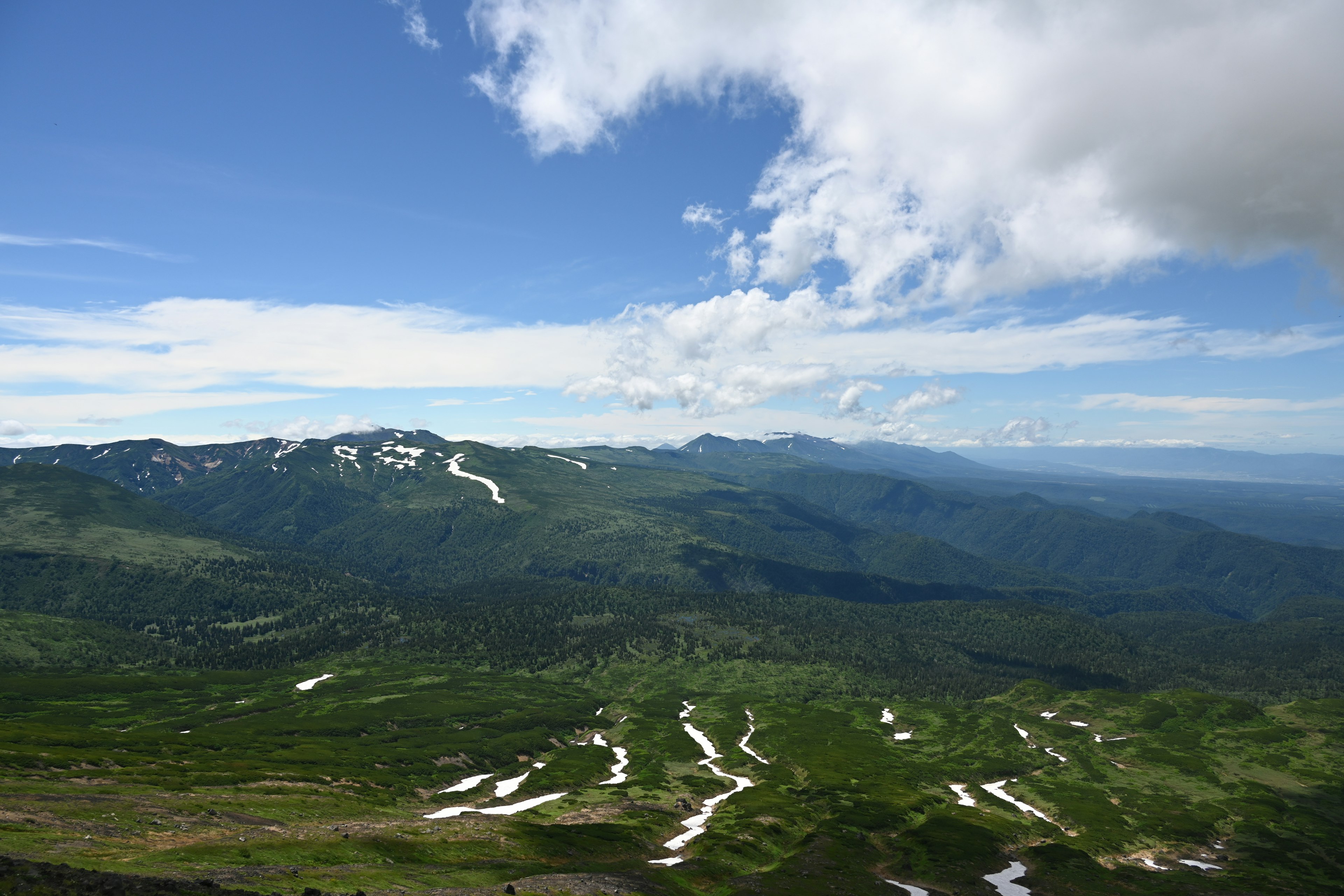 Vista expansiva de montañas verdes con parches de nieve bajo un cielo azul y nubes blancas