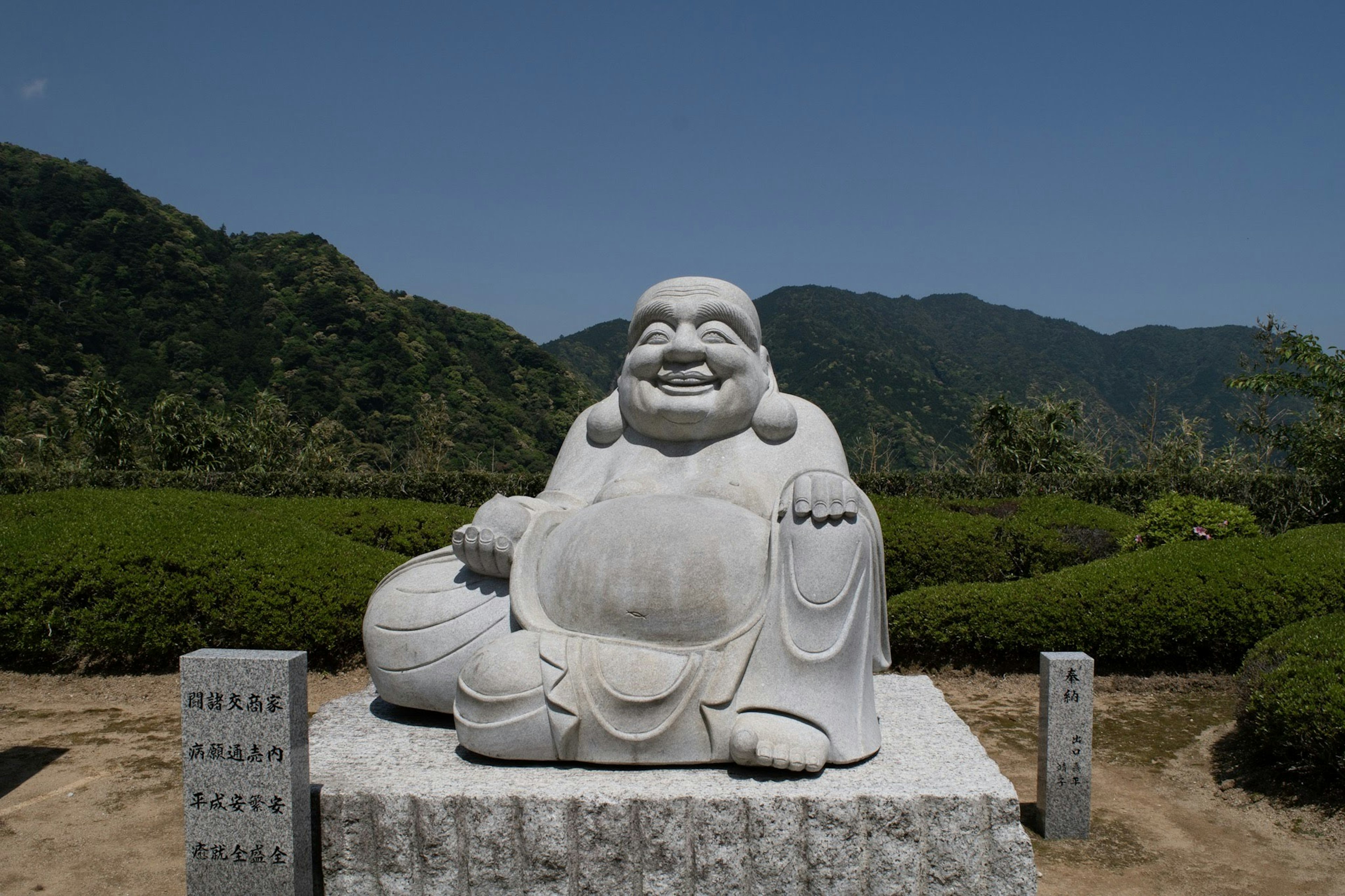 A white statue of a laughing Buddha seated against a mountainous background