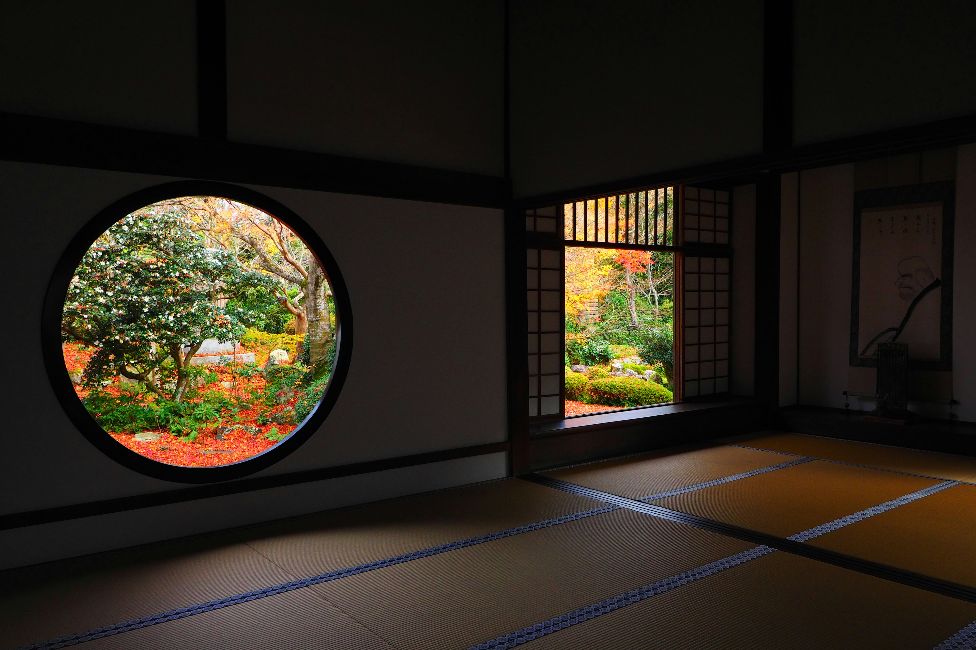 Interior of a traditional Japanese room featuring a circular window with a beautiful garden view and a square window showing colorful trees