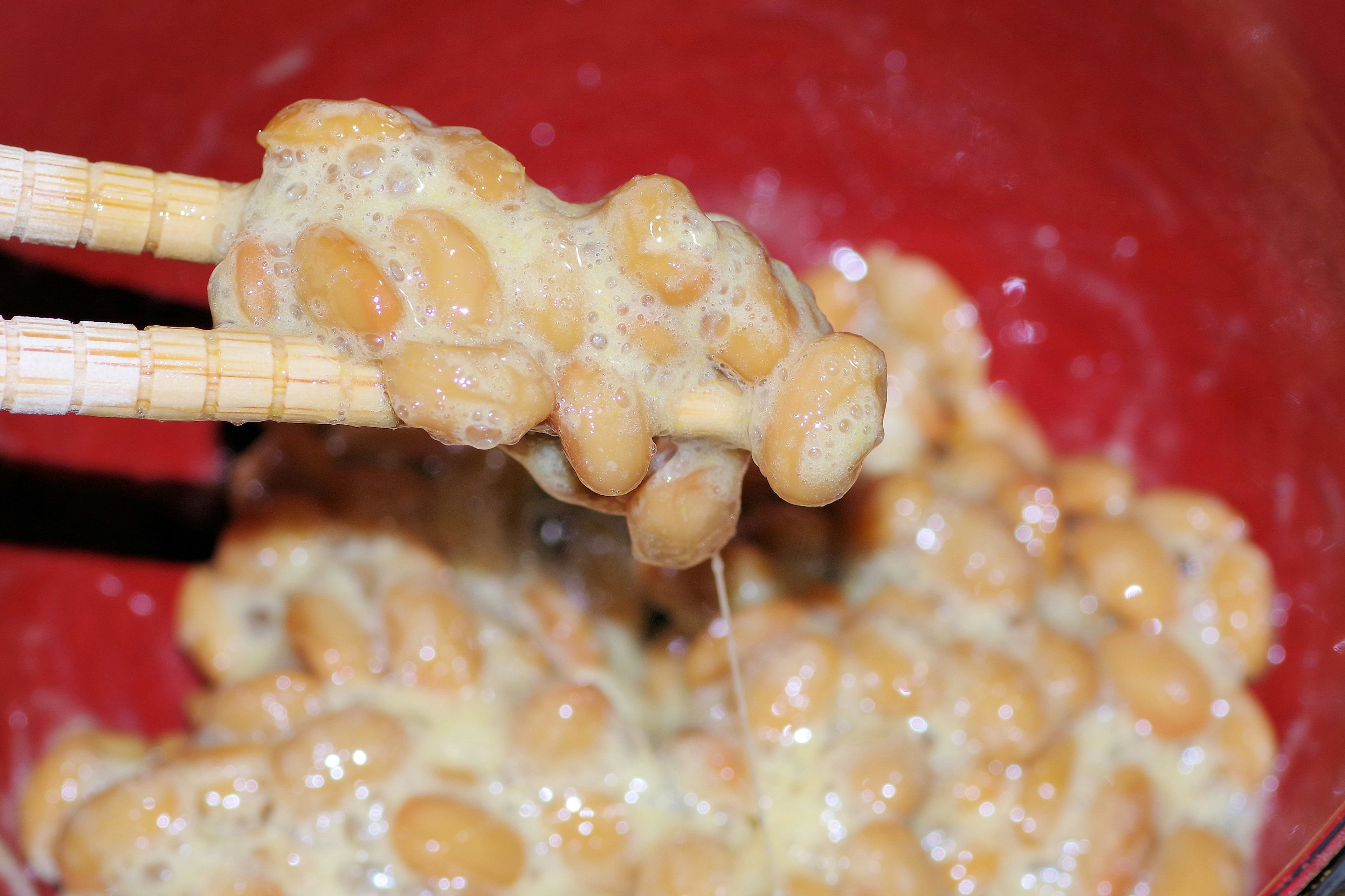 Natto being lifted with chopsticks sticky texture in a red bowl