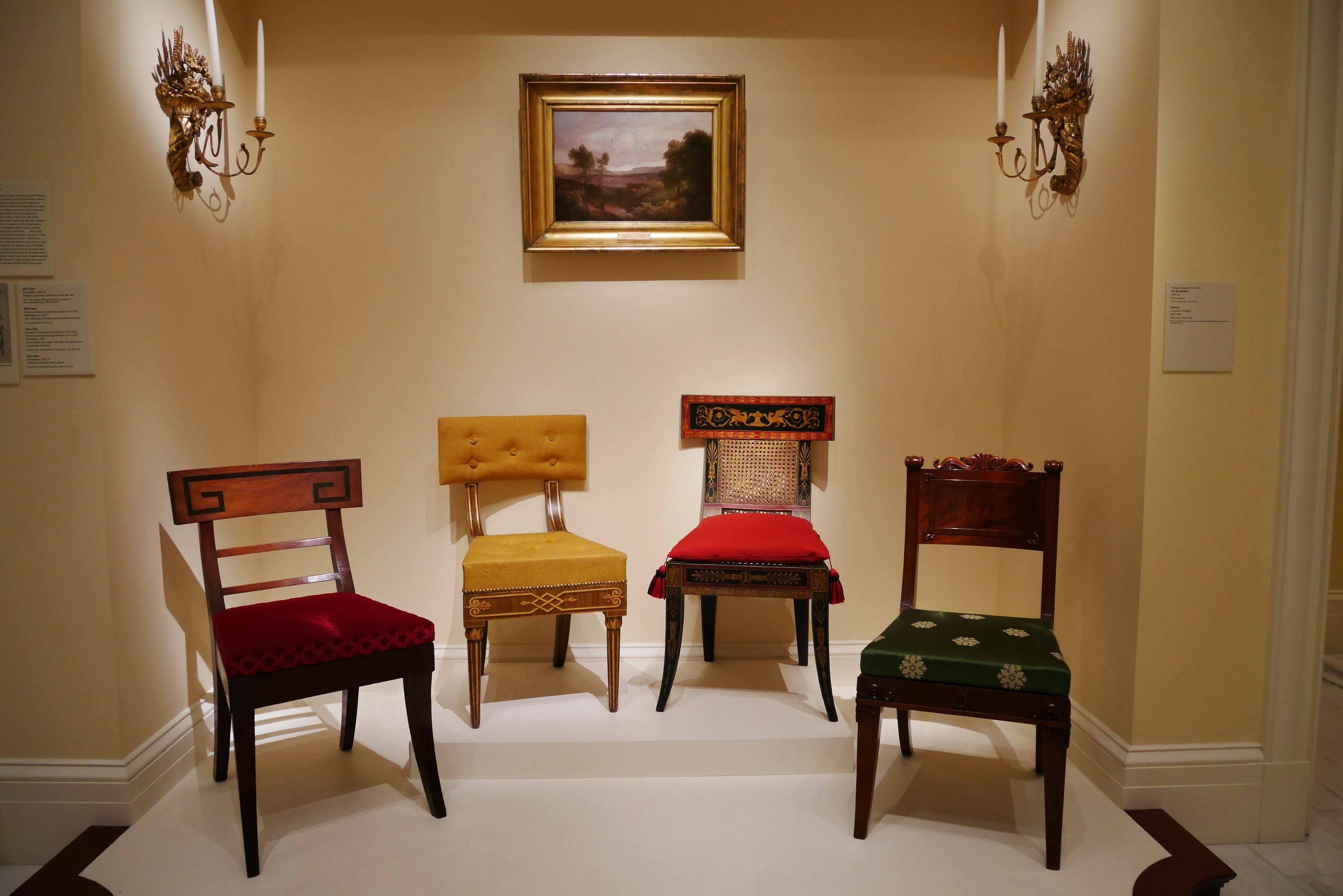 Four different styled chairs arranged in a corner of a room with a painting on the wall