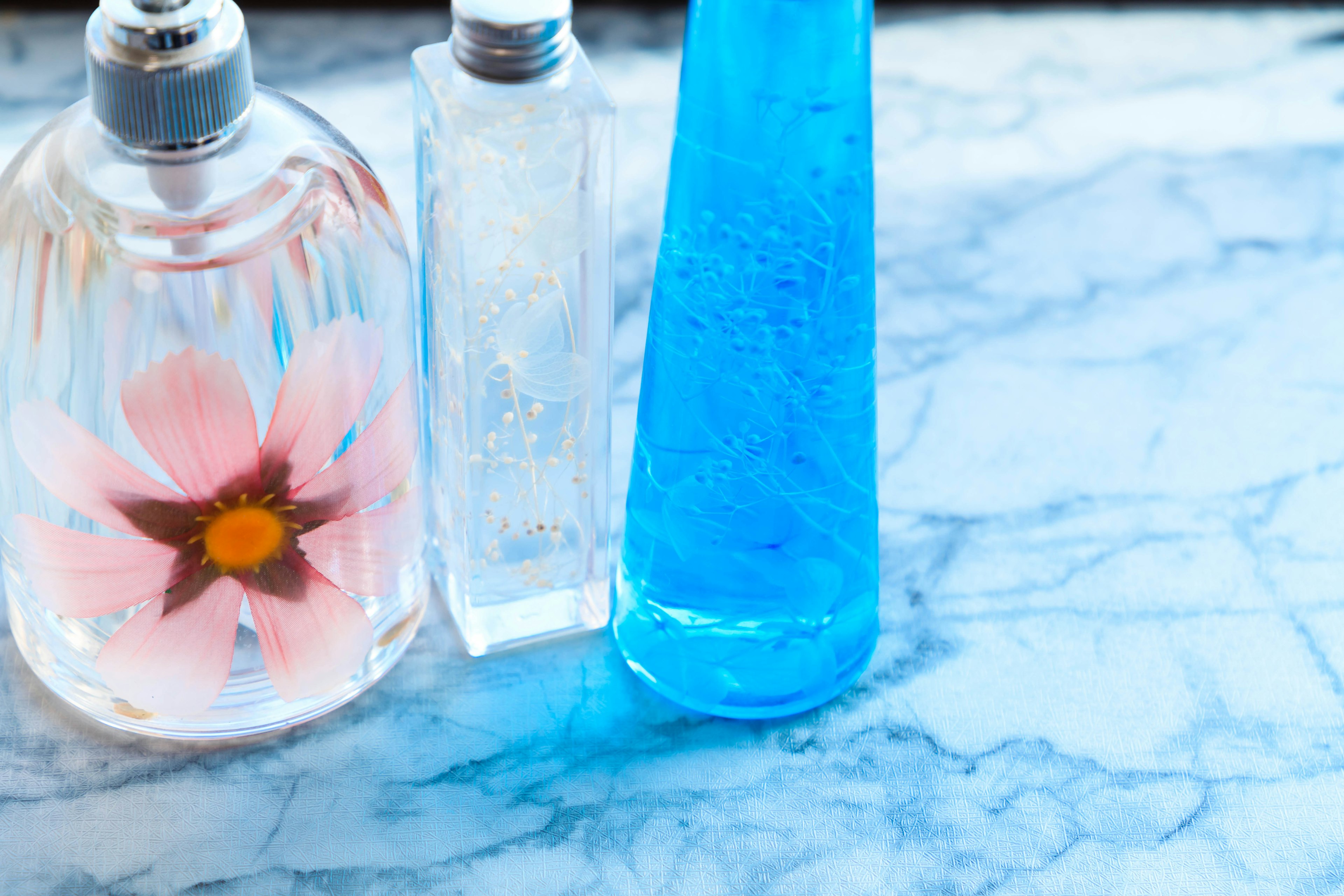 Tres botellas decorativas incluyendo una botella de perfume floral y una botella de líquido azul sobre una superficie de mármol