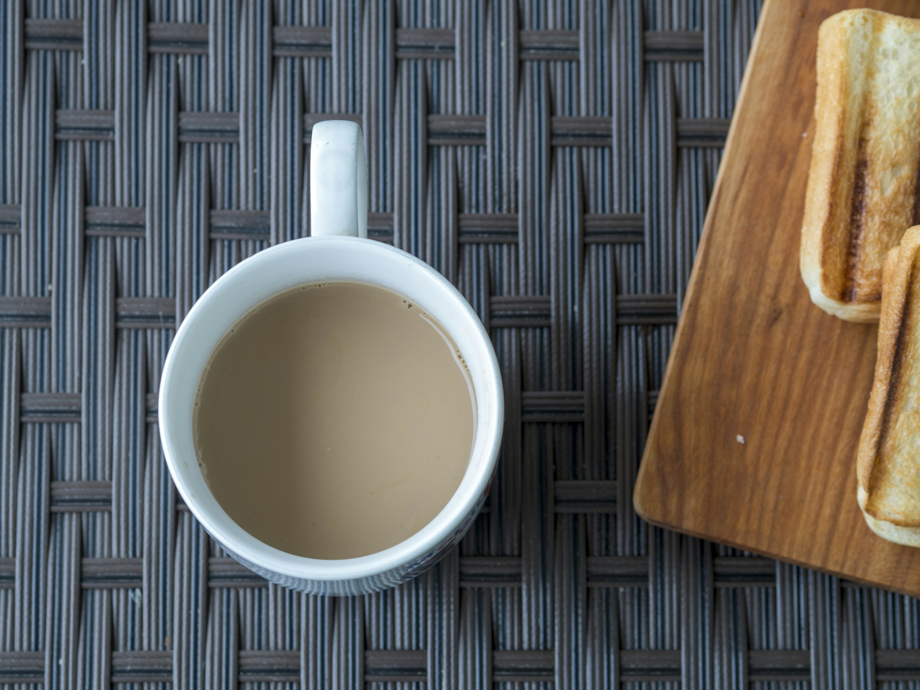 Una taza blanca con café claro al lado de tostadas en una bandeja de madera