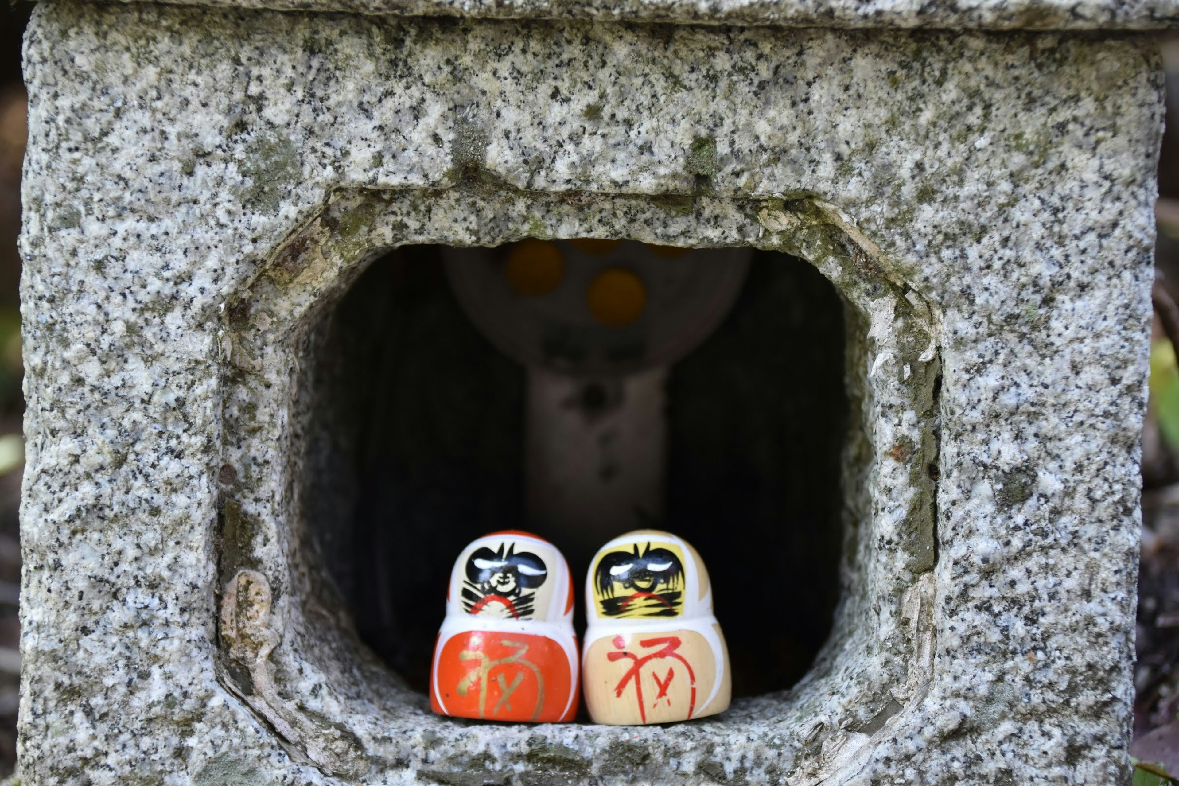 Dos muñecos Daruma dentro de una linterna de piedra con una estatua al fondo