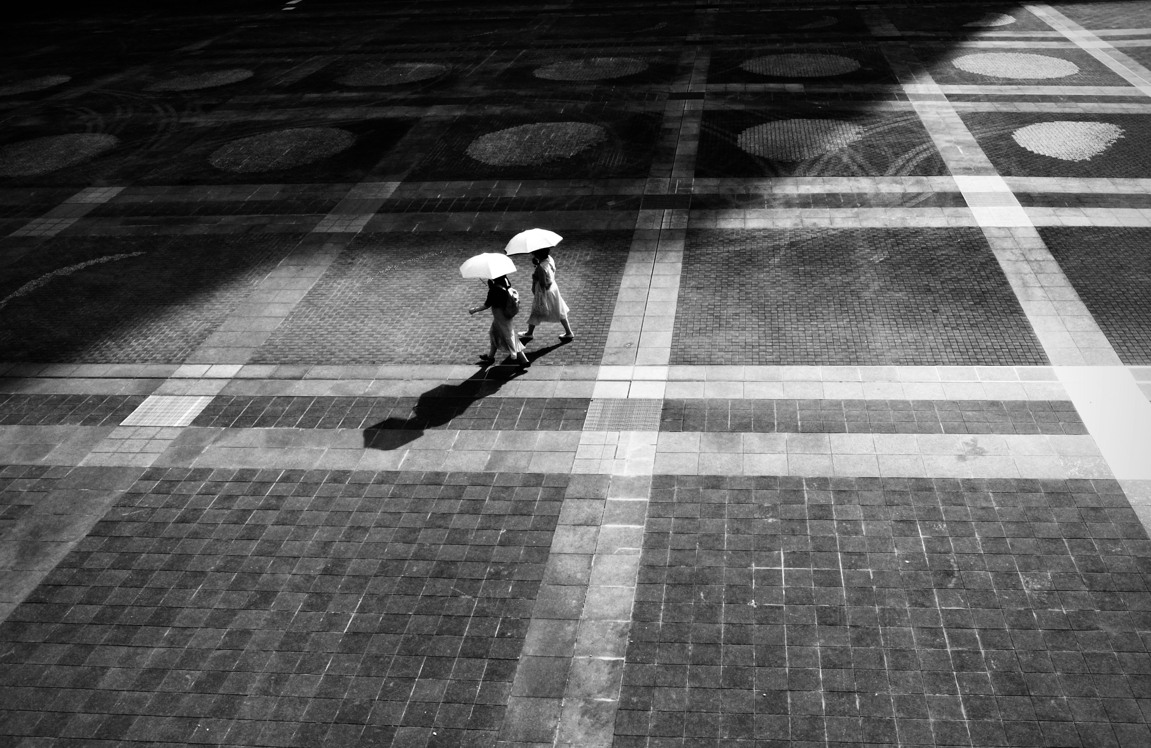 Escena urbana en blanco y negro con dos personas caminando sobre un pavimento de baldosas sosteniendo paraguas