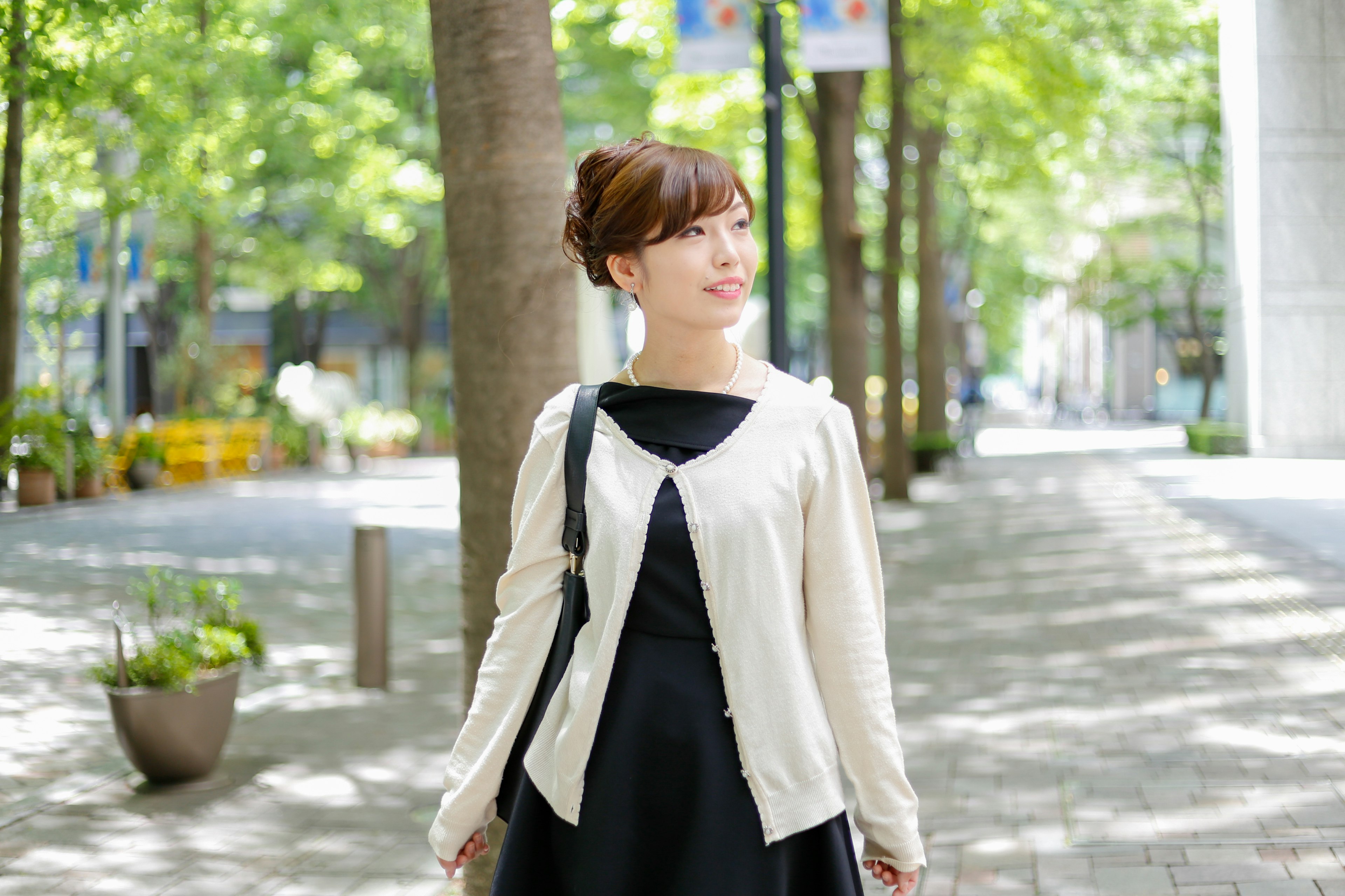 A smiling woman in a city street wearing a white cardigan with green trees lining the walkway