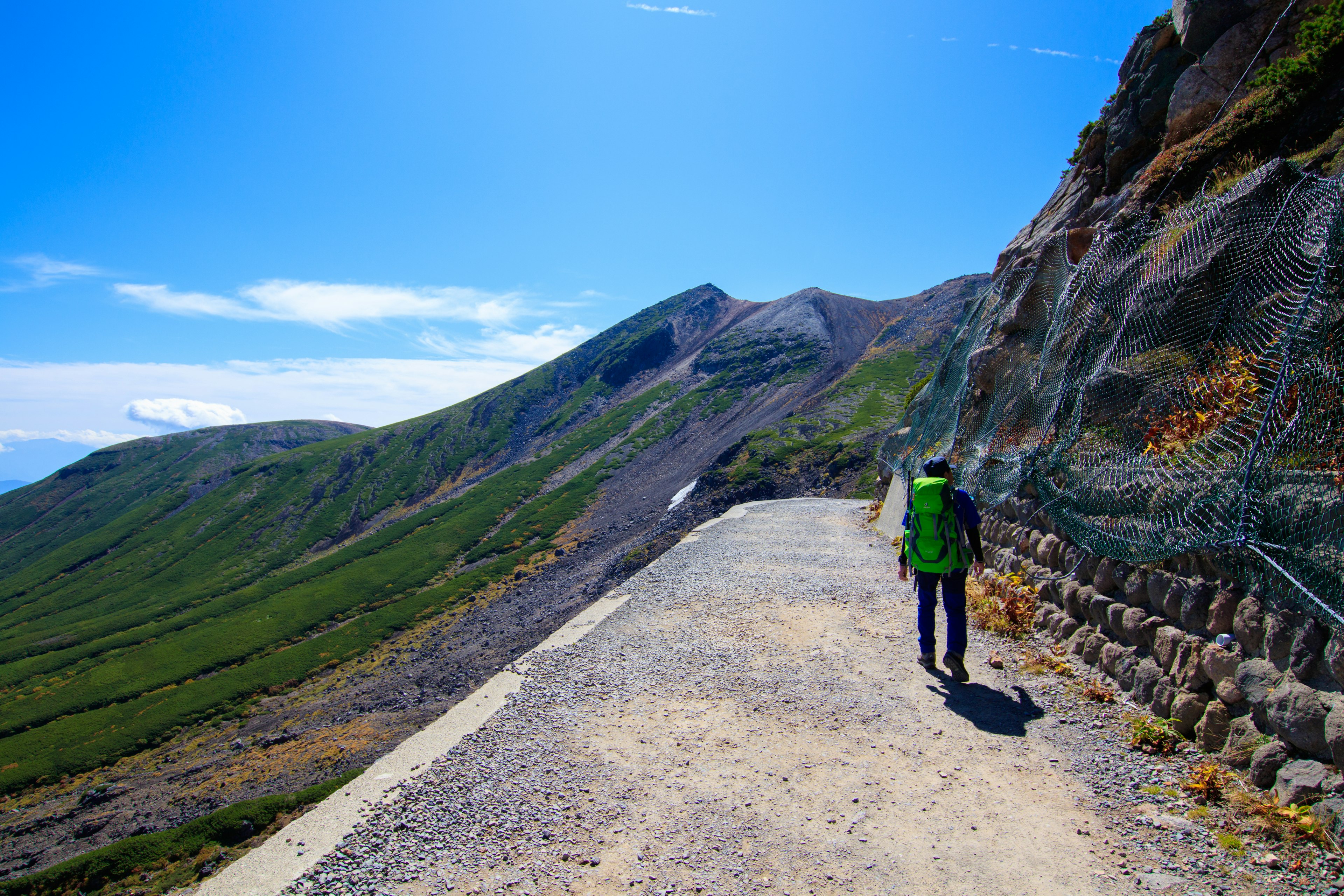 Ein Wanderer, der entlang eines malerischen Bergpfades mit üppigem Grün und blauem Himmel geht