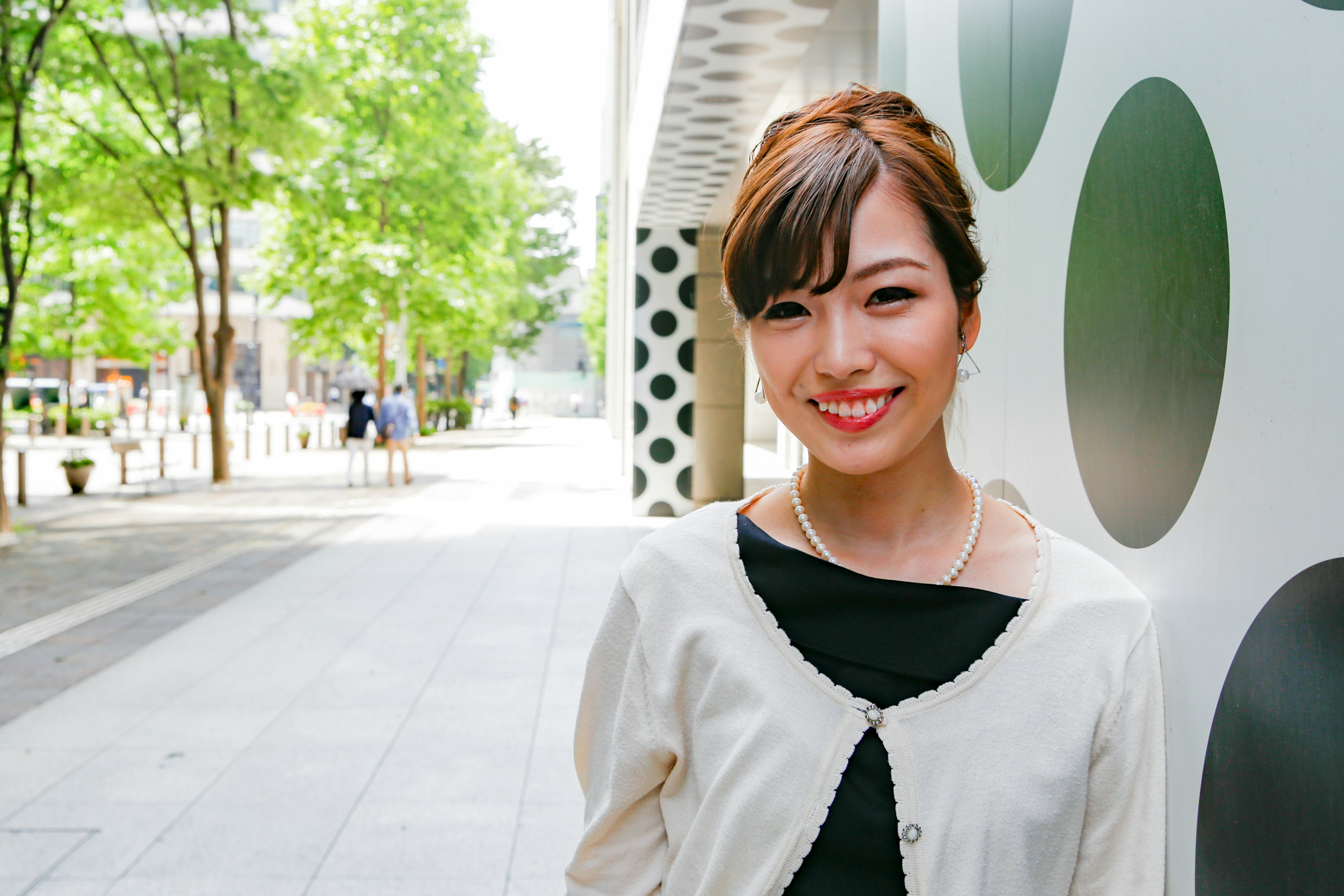 Smiling woman in a park wearing a white jacket surrounded by green trees and circular walls