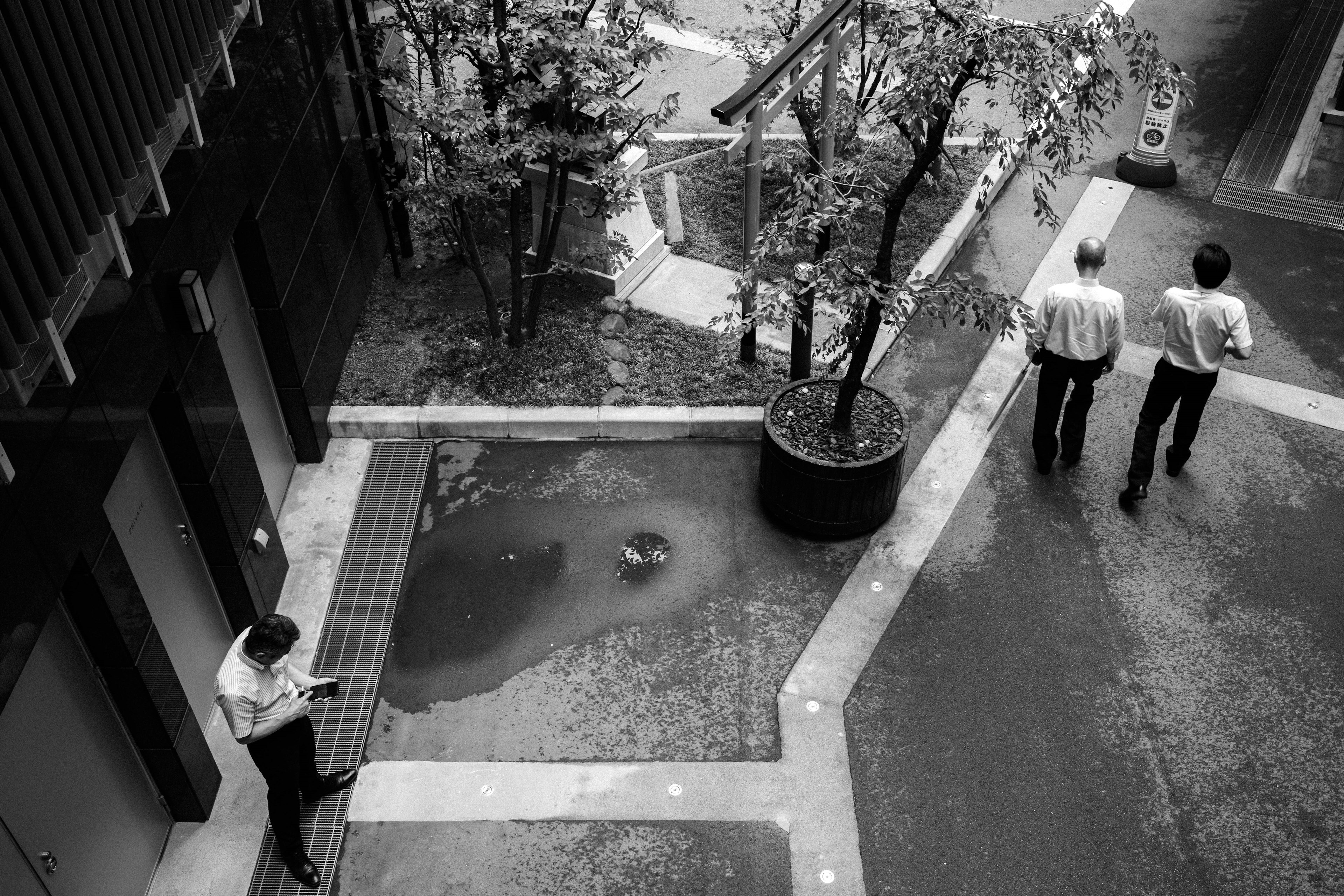 Scène urbaine en noir et blanc avec deux hommes d'affaires marchant et une personne assise sur un banc utilisant un smartphone