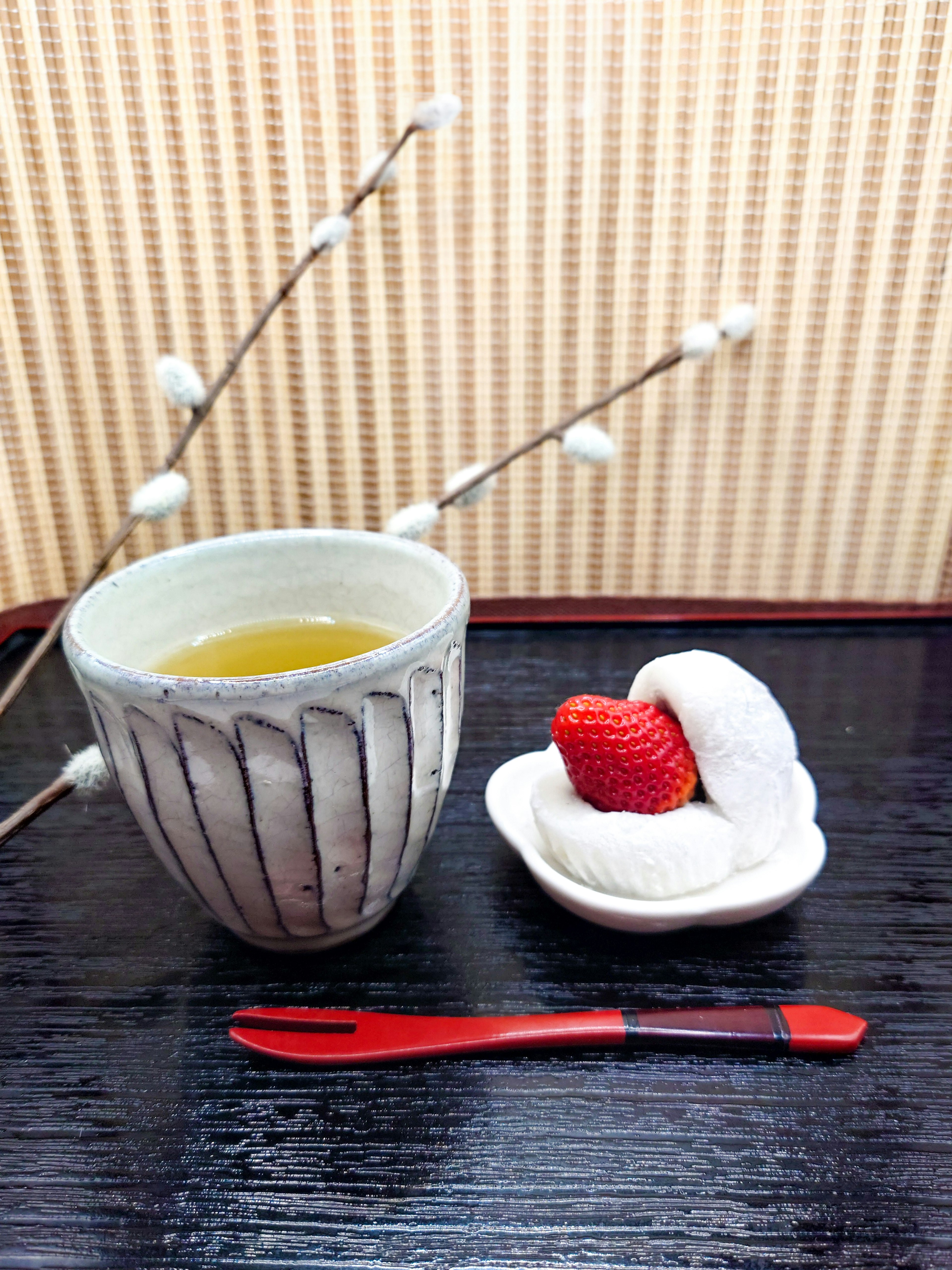 A cup of green tea beside a strawberry daifuku on a black tray