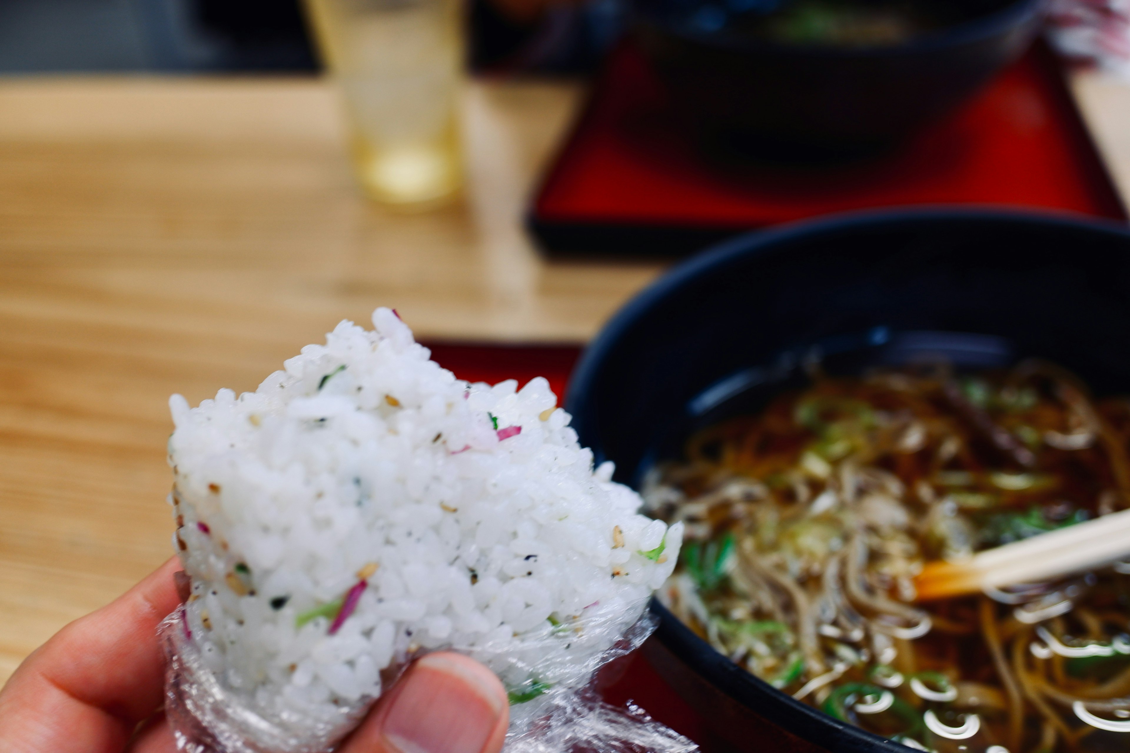 A hand holding a rice ball with a bowl of noodles in the background