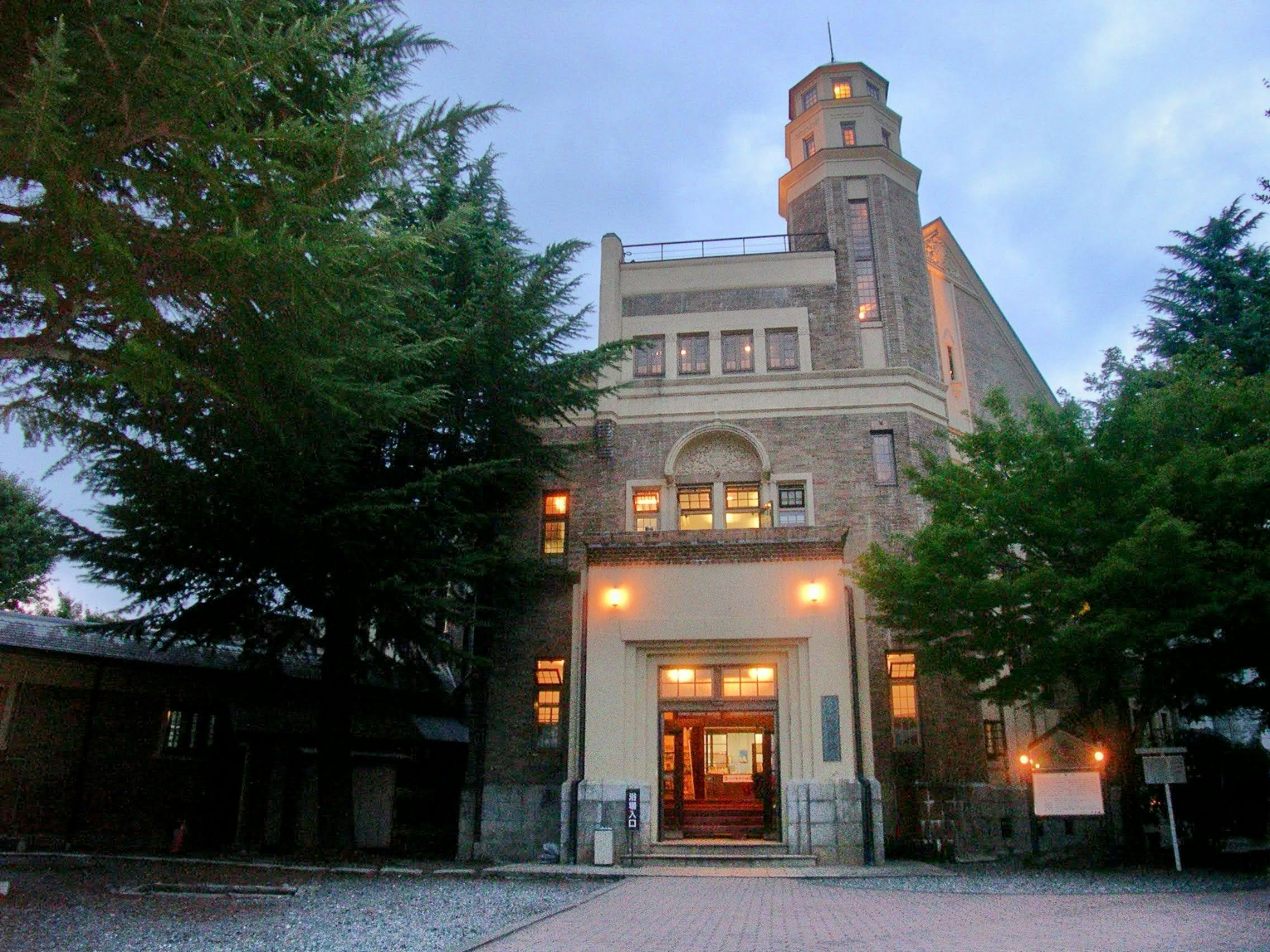 Historic building exterior illuminated at dusk surrounded by green trees featuring a distinctive tower