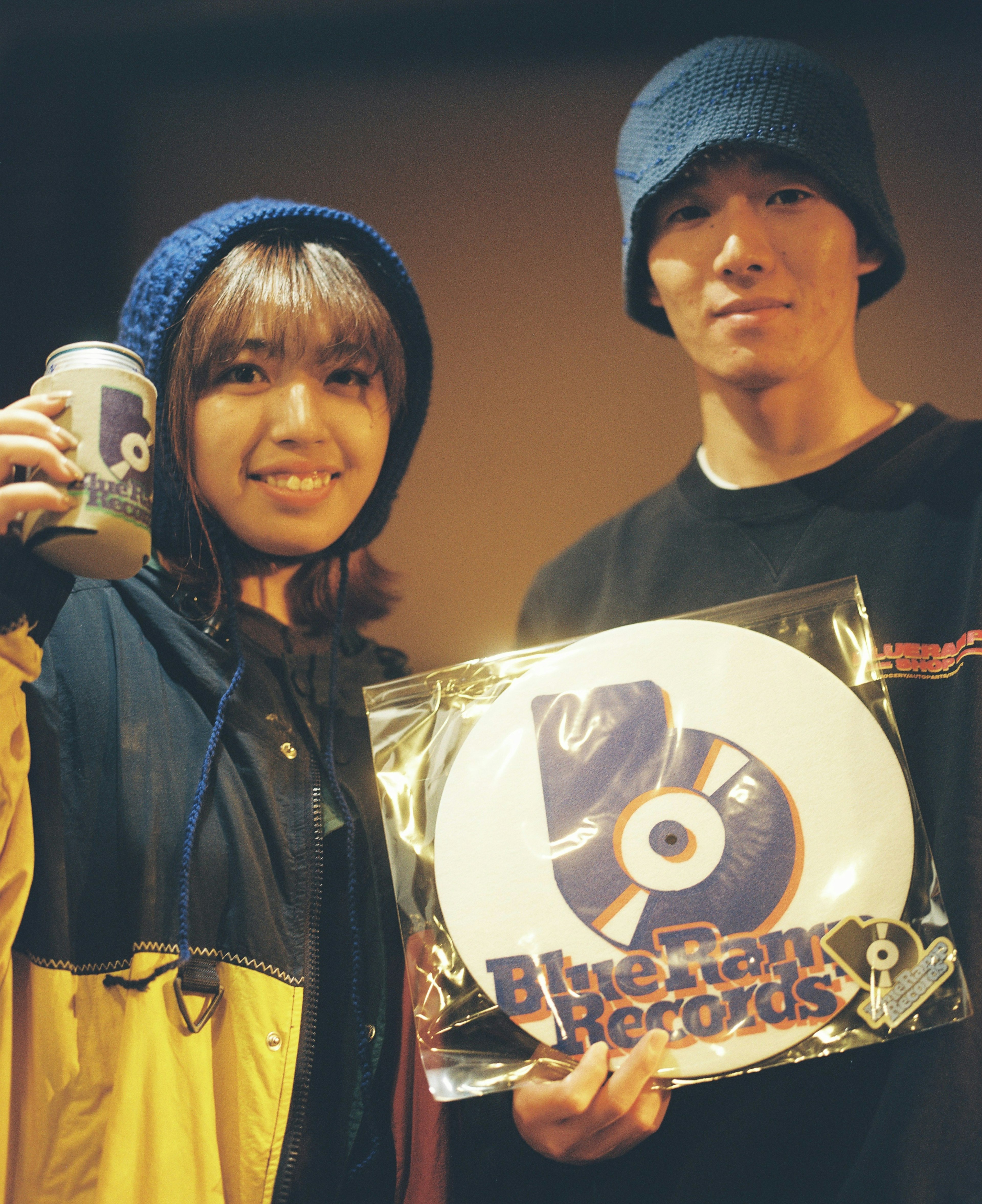 Joven hombre y mujer con sombreros azules sosteniendo una placa del logo de Blue Dunk Records y una bebida