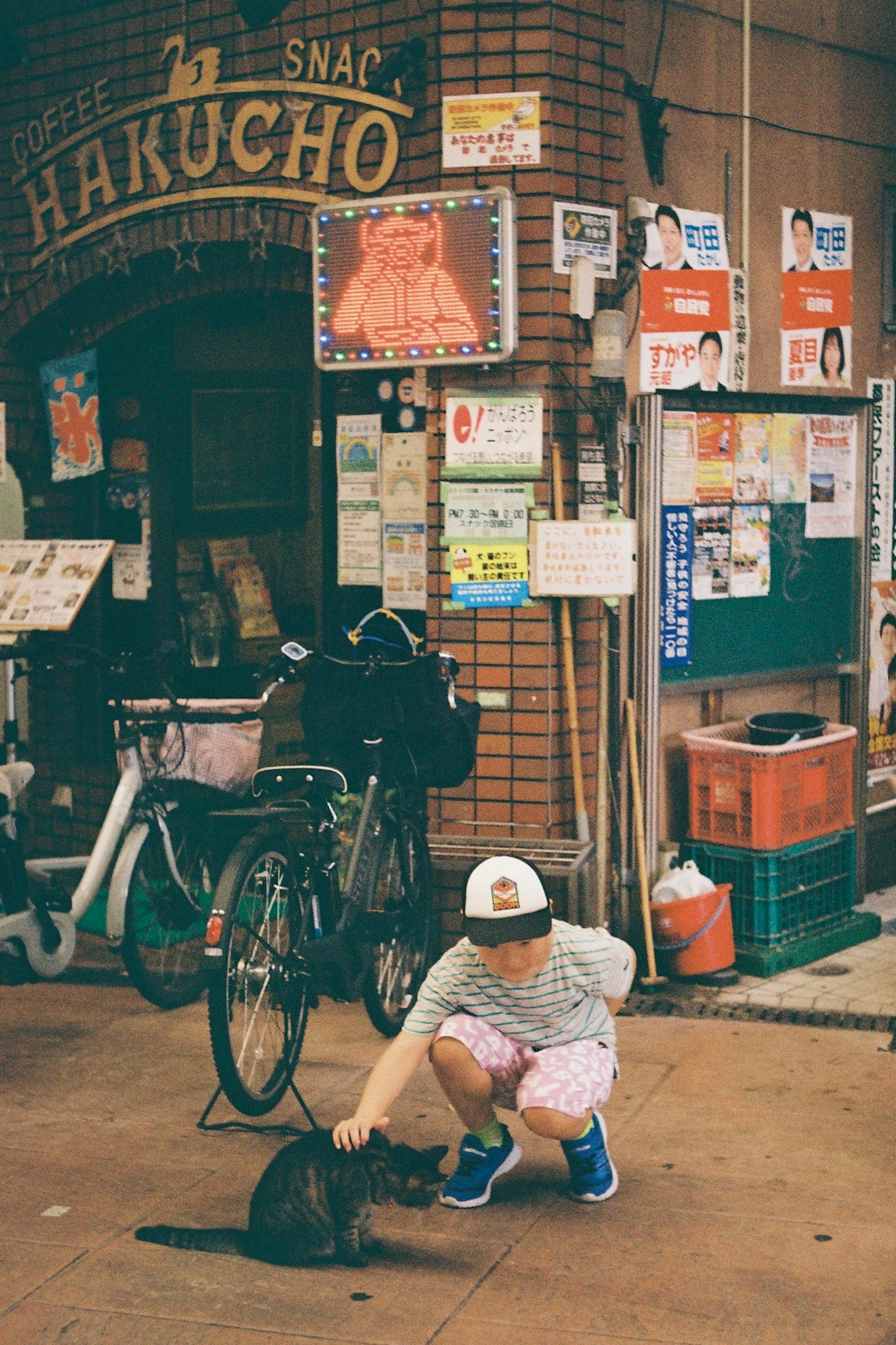 一个孩子在HAKUCHO小吃店前抚摸一只黑猫，旁边有自行车