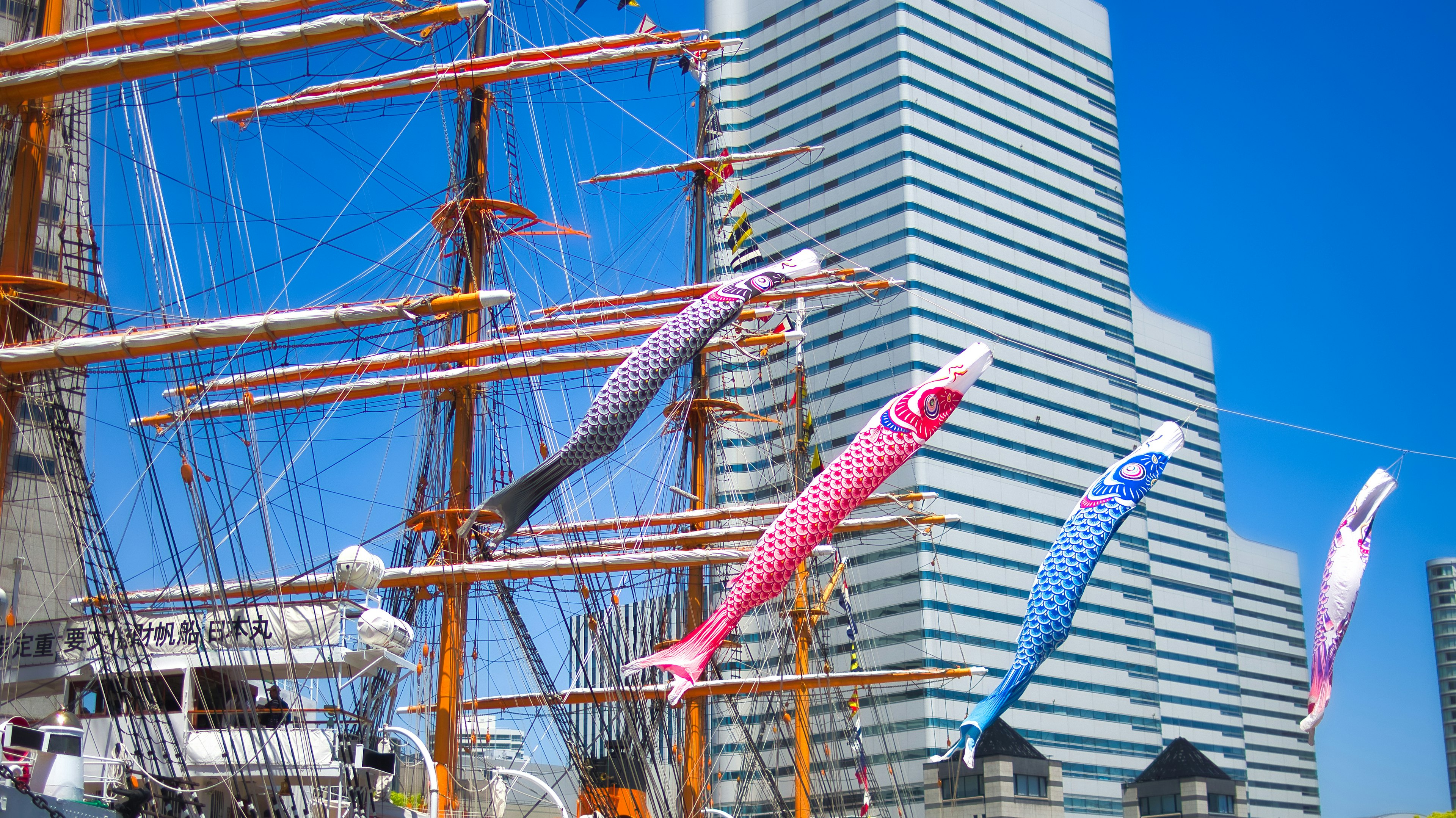 A scene featuring a sailing ship and a modern building under a blue sky with koi flags fluttering