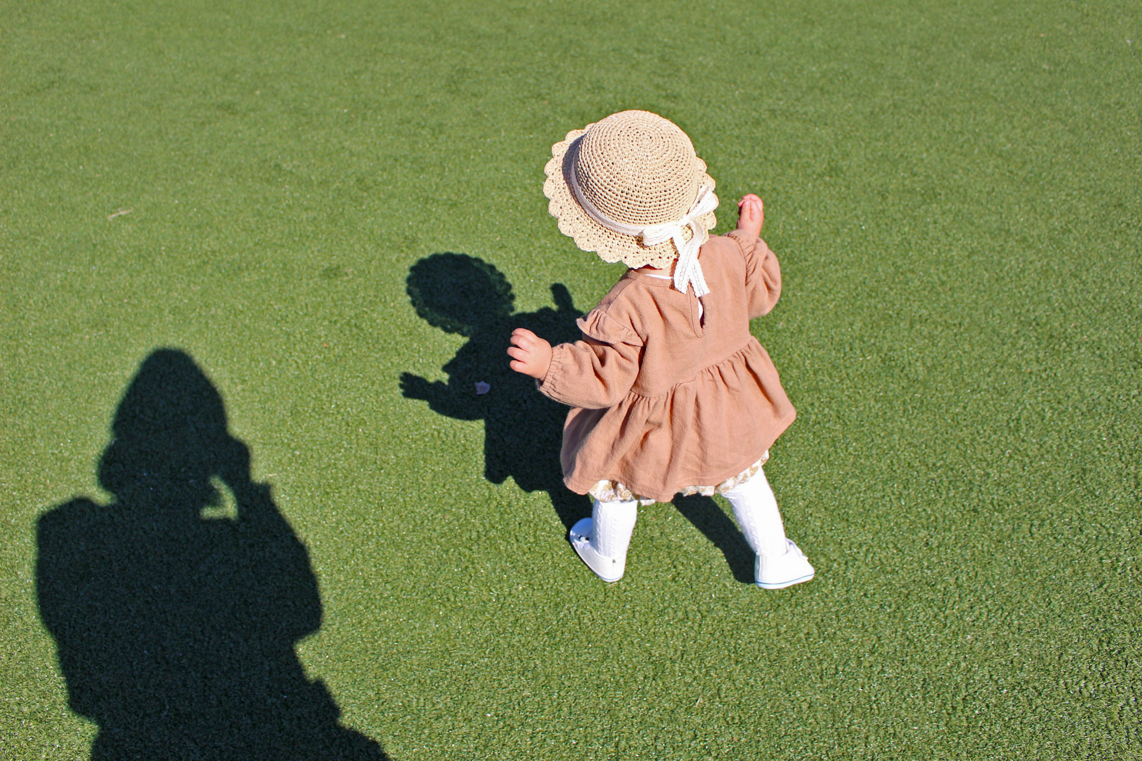 Un enfant en robe brune et chapeau jouant sur de l'herbe verte avec une ombre