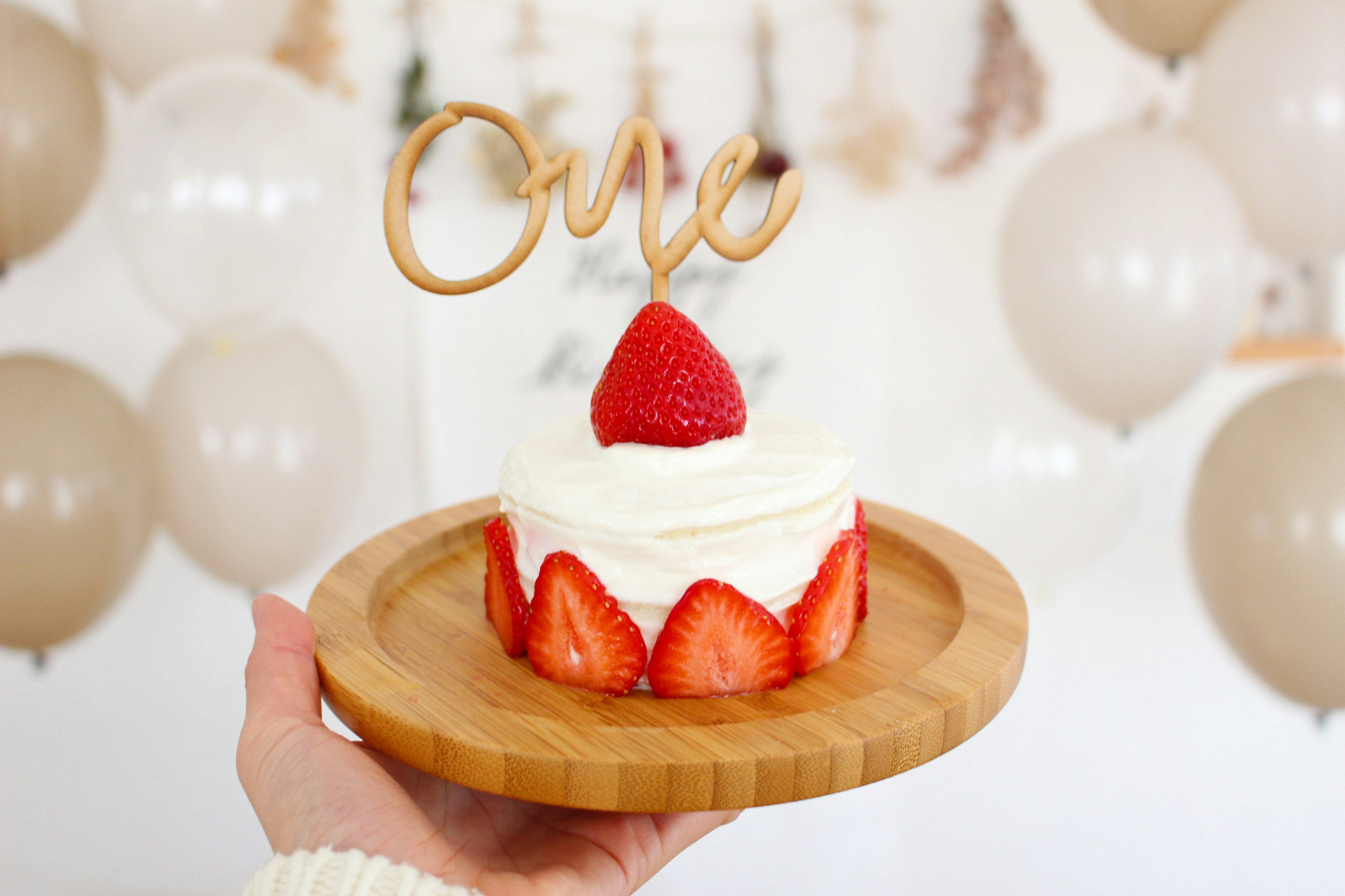 Una torta di fragole con una decorazione in legno 'One' circondata da palloni beige