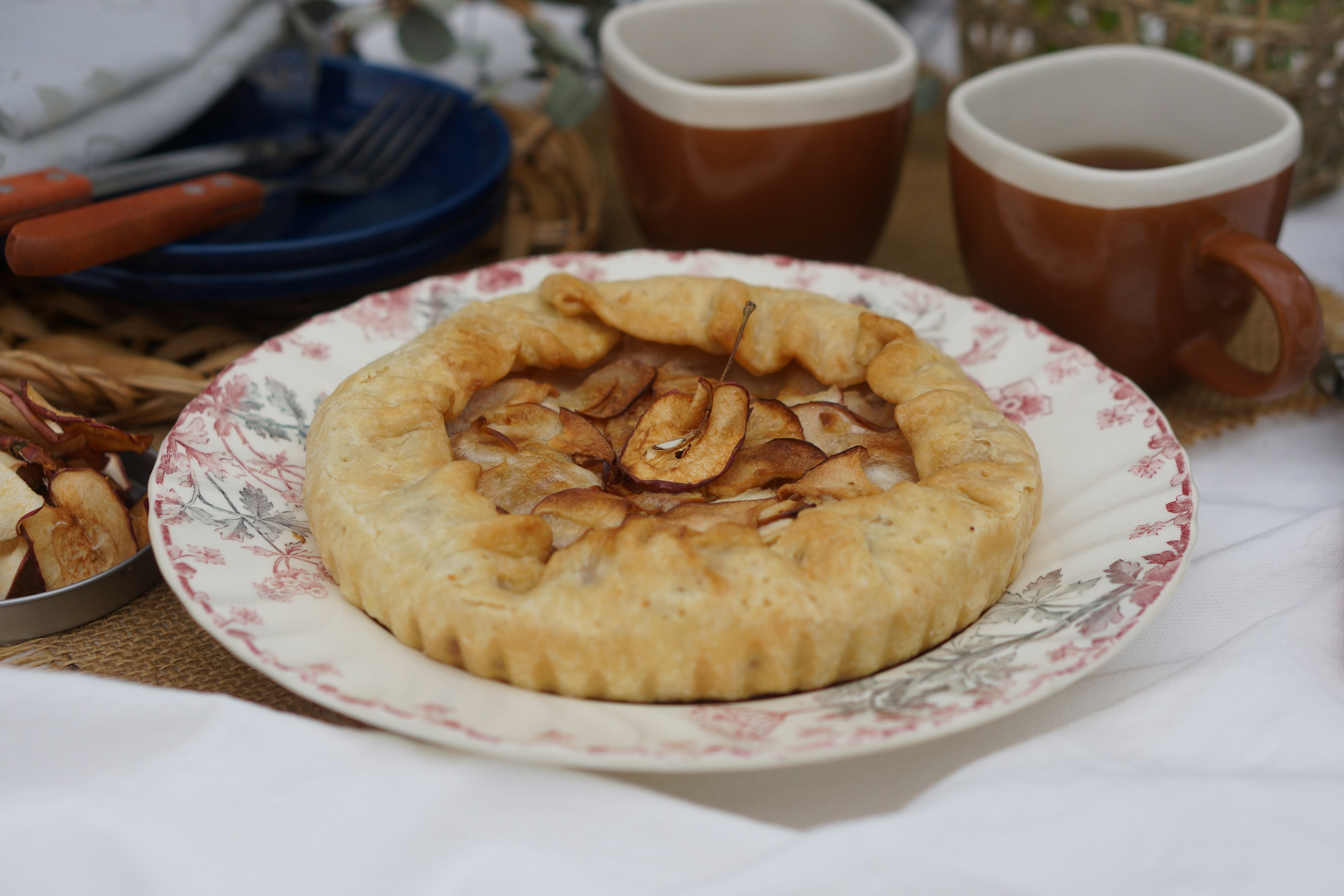 Apfelkuchen auf einem blumigen Teller mit zwei Kaffeetassen im Hintergrund