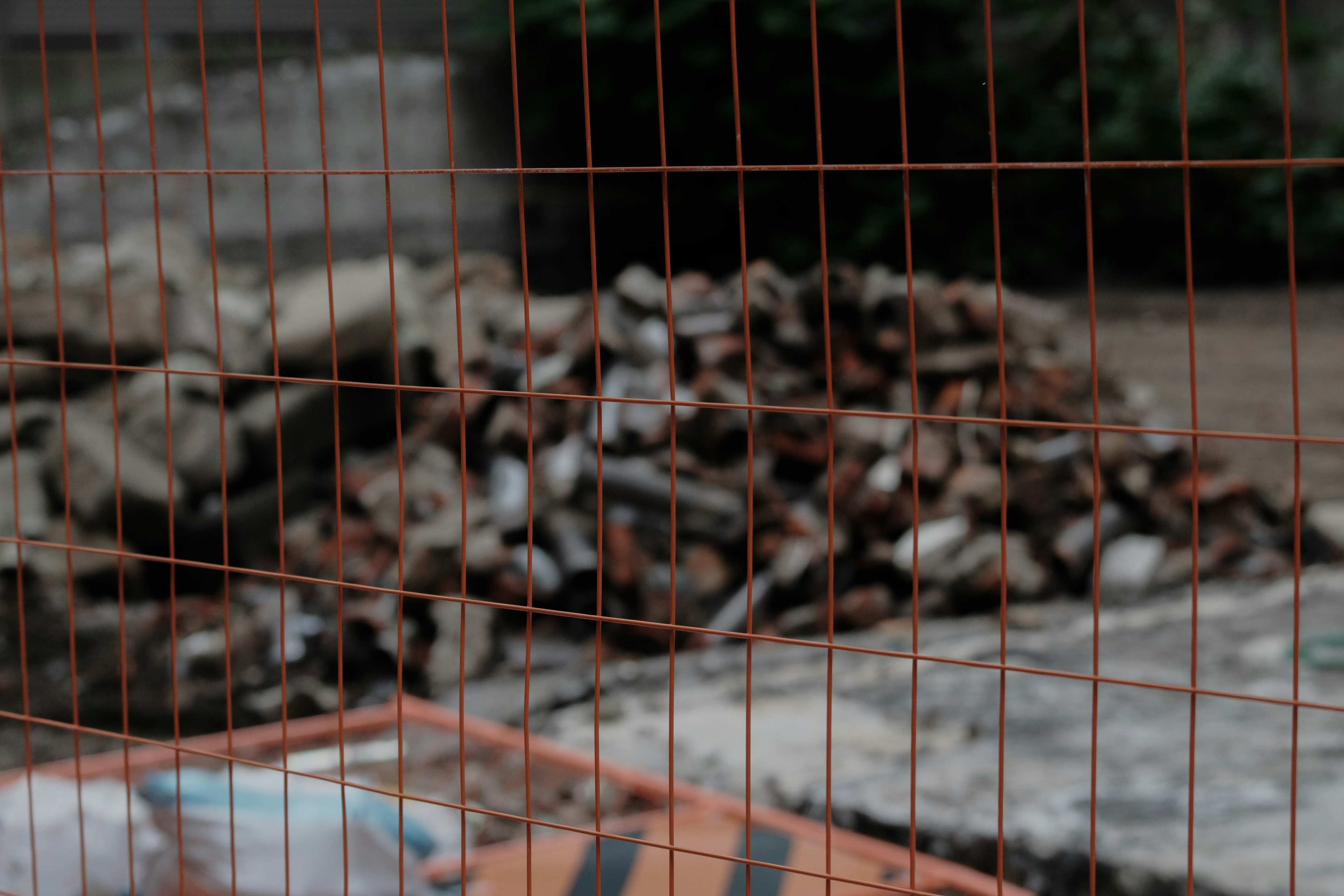 A pile of wood seen through an orange mesh fence with a concrete surface