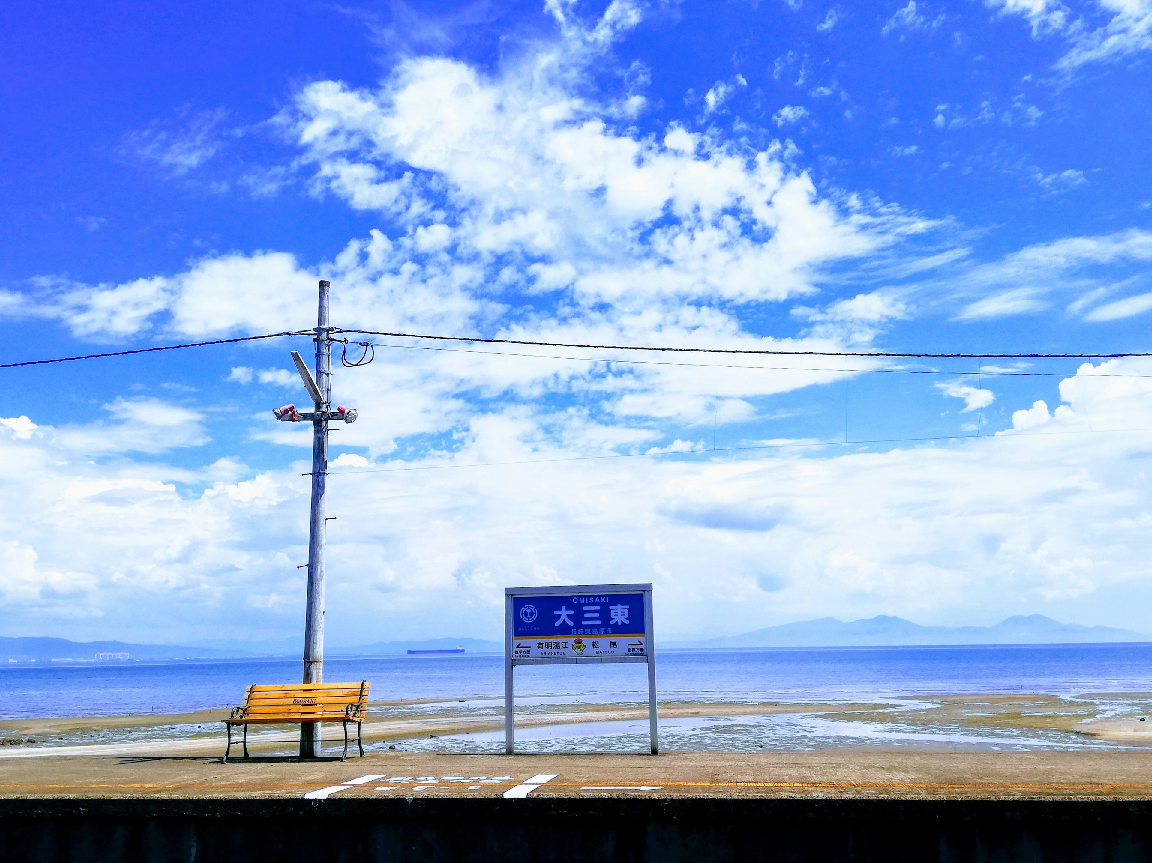 青空と雲が広がる海岸の風景にベンチと標識がある