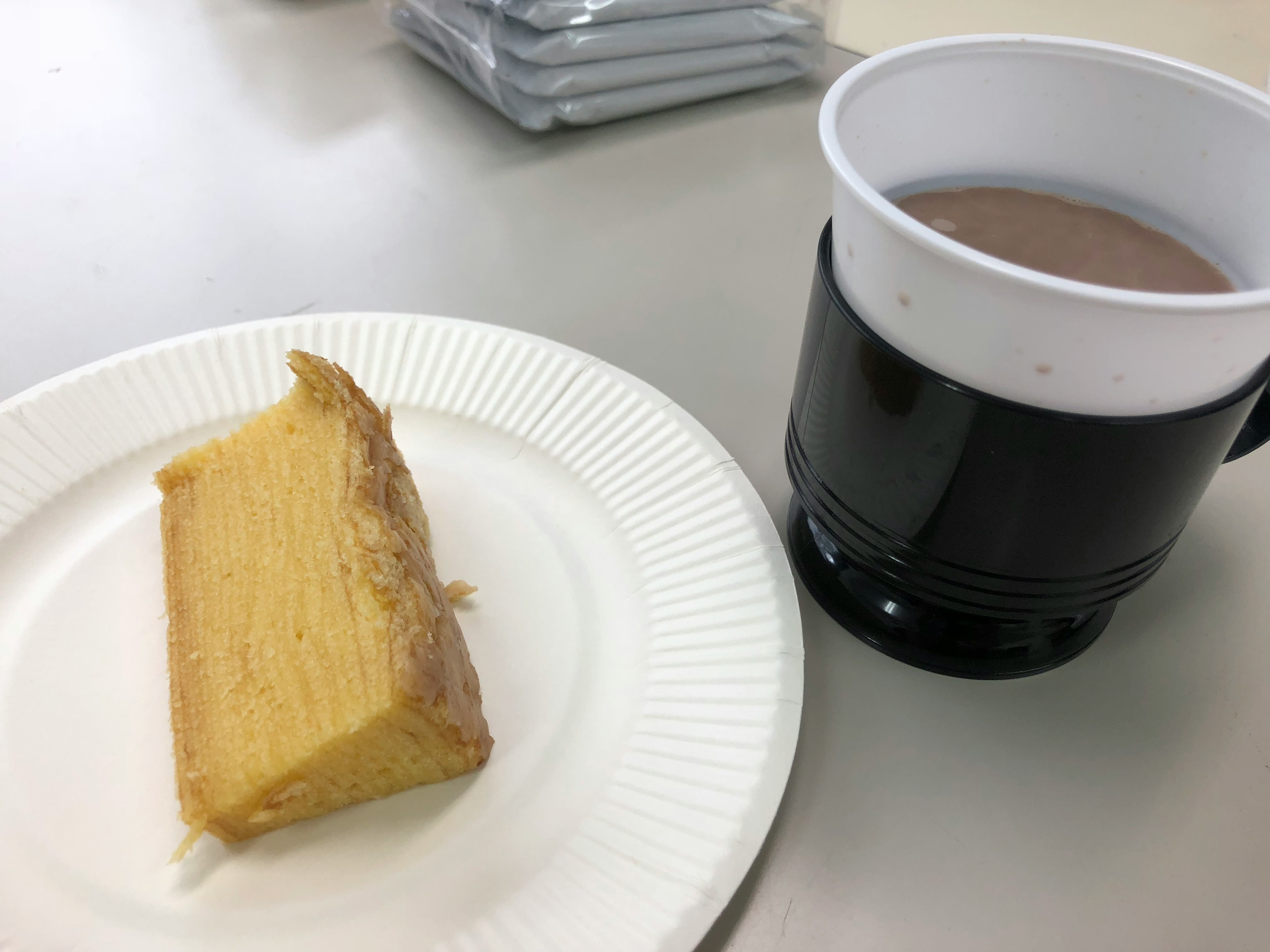 Una rebanada de pastel amarillo en un plato blanco junto a una taza negra de bebida