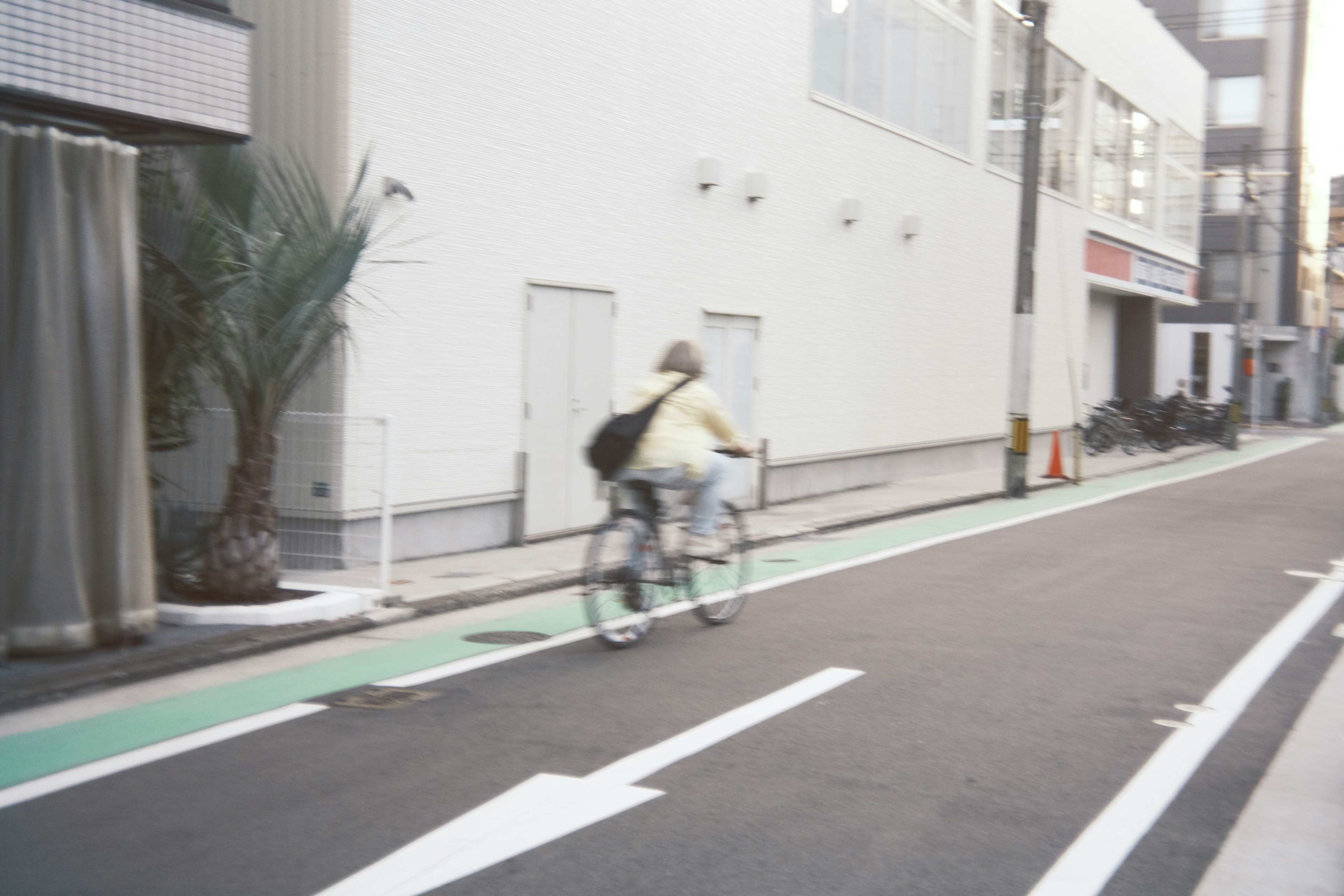 Una mujer montando una bicicleta en una calle urbana con un edificio blanco y una ciclovía verde al fondo