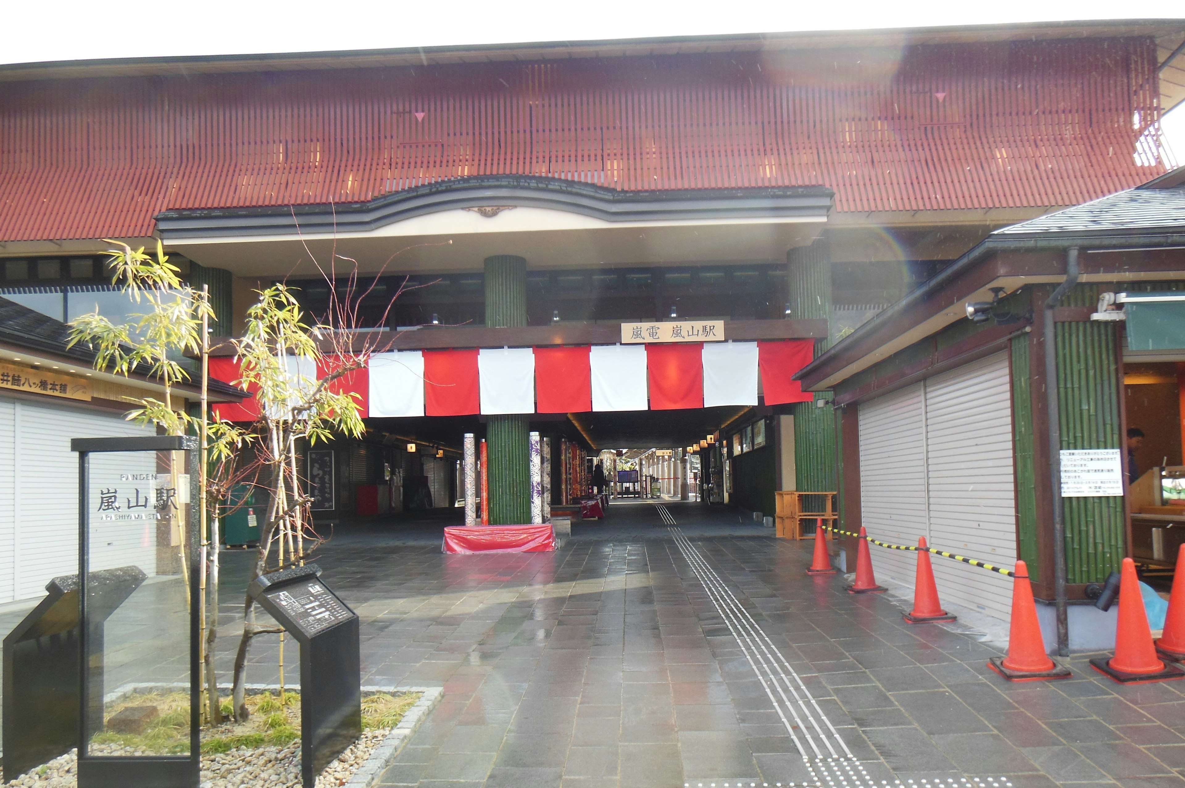 Entrada de un establecimiento comercial con un techo rojo y una bandera blanca y roja rodeada de conos naranjas