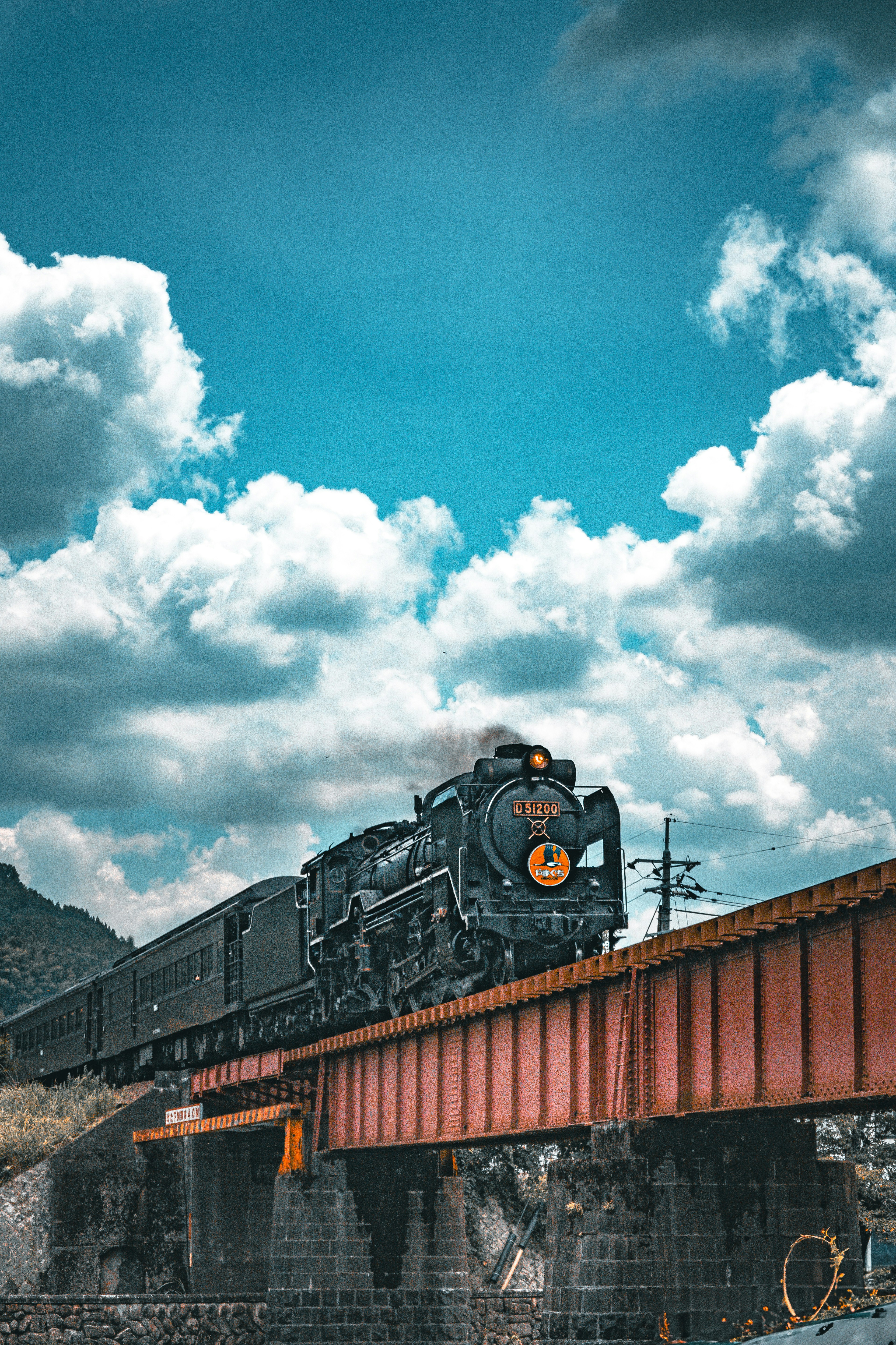 Locomotive à vapeur traversant un pont rouge sous un ciel bleu avec des nuages blancs