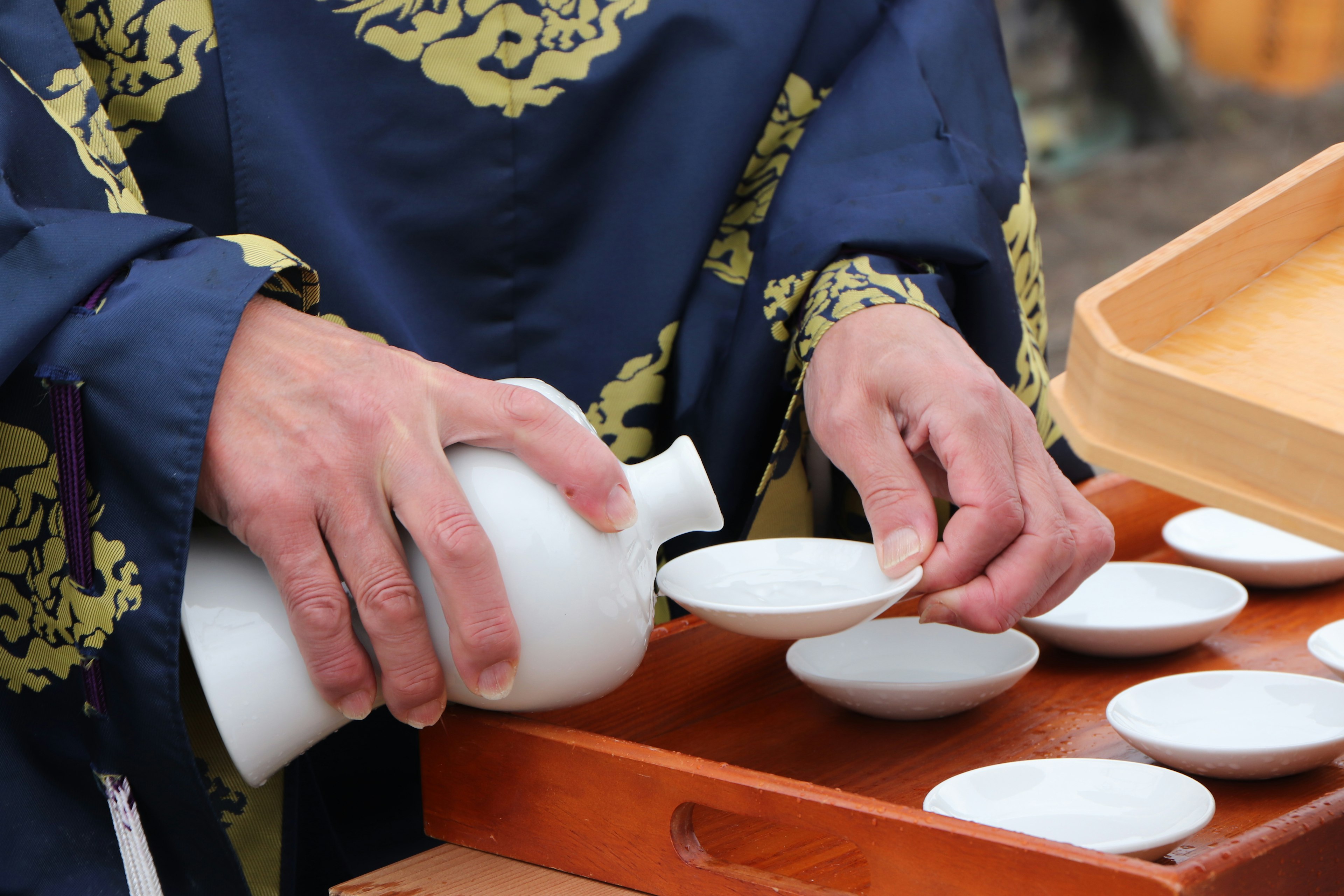 A person in traditional attire pouring from a white ceramic vessel into small dishes