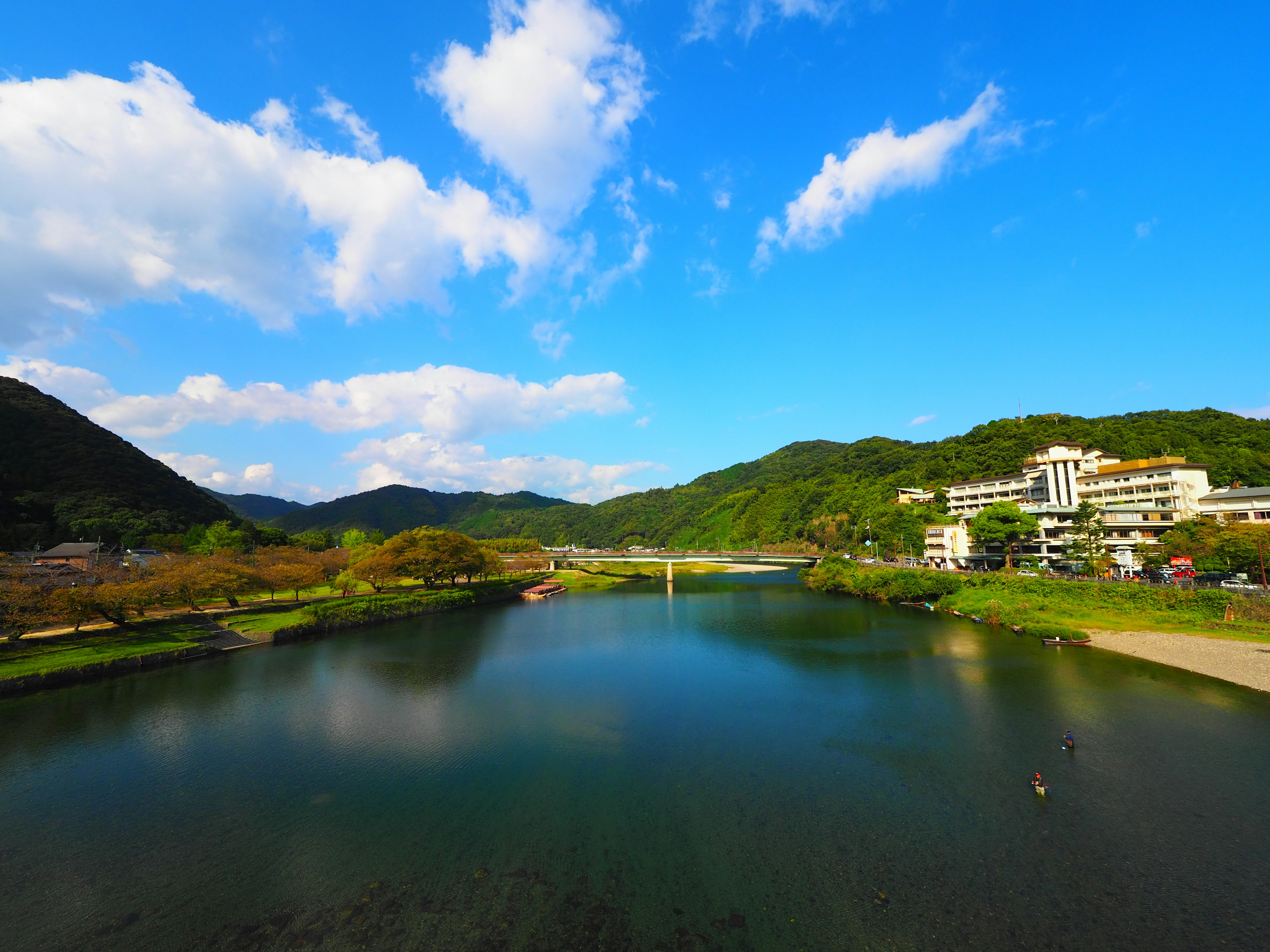 Malersiche Aussicht auf einen ruhigen Fluss, der den blauen Himmel und die weißen Wolken widerspiegelt, umgeben von üppigen grünen Bergen