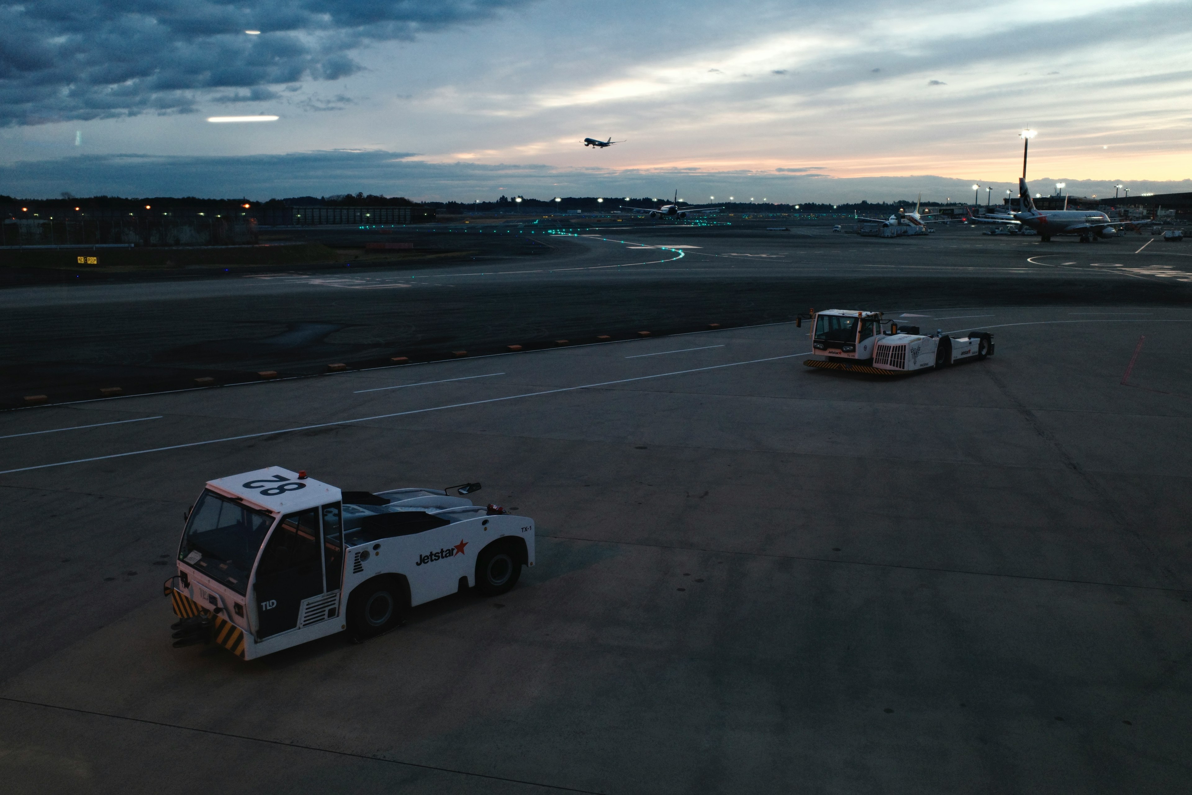 Two ground support vehicles on an airport runway with a plane taking off in the background