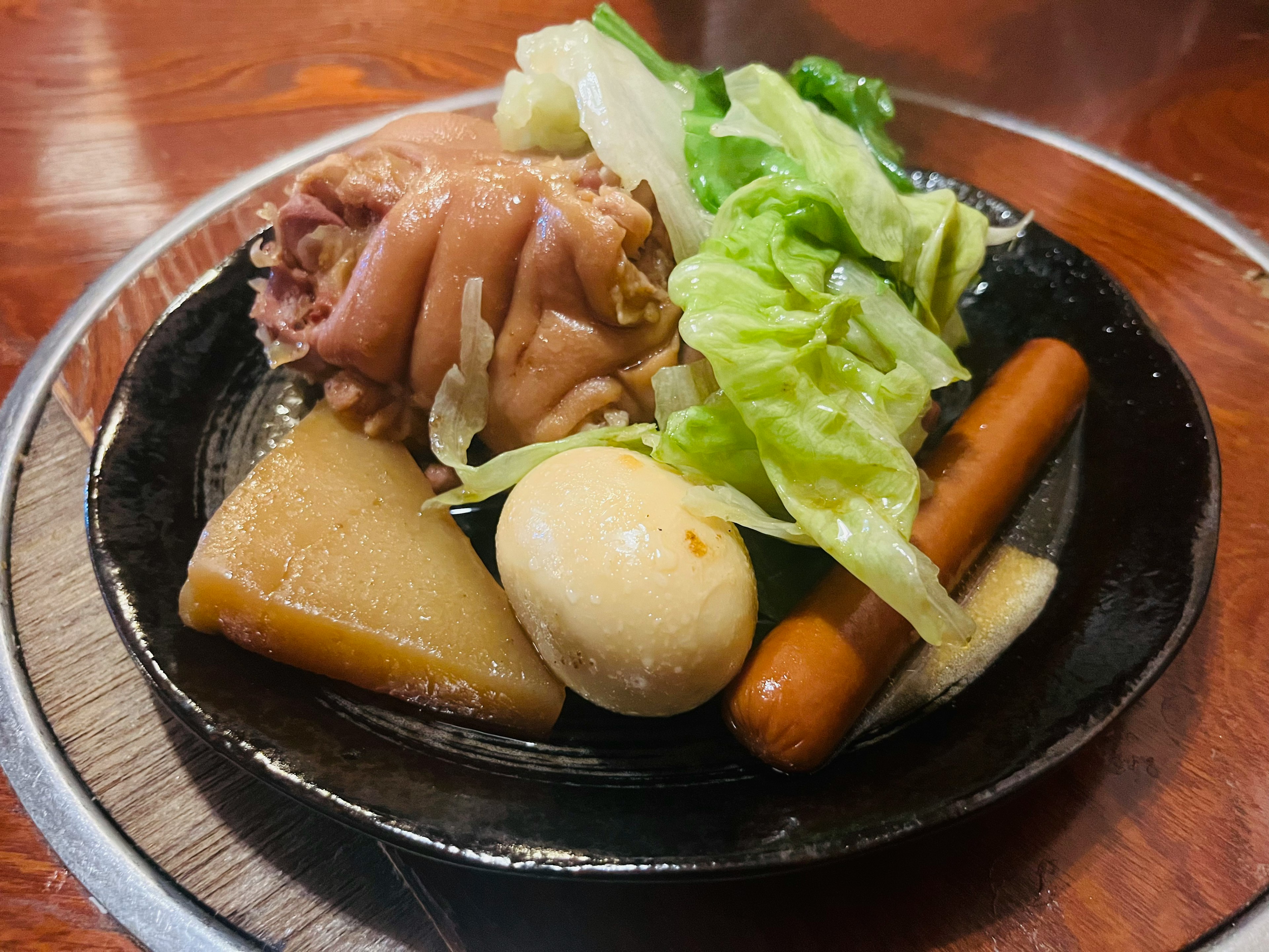 A plate of simmered dish featuring chicken, boiled egg, cabbage, daikon, and sausage