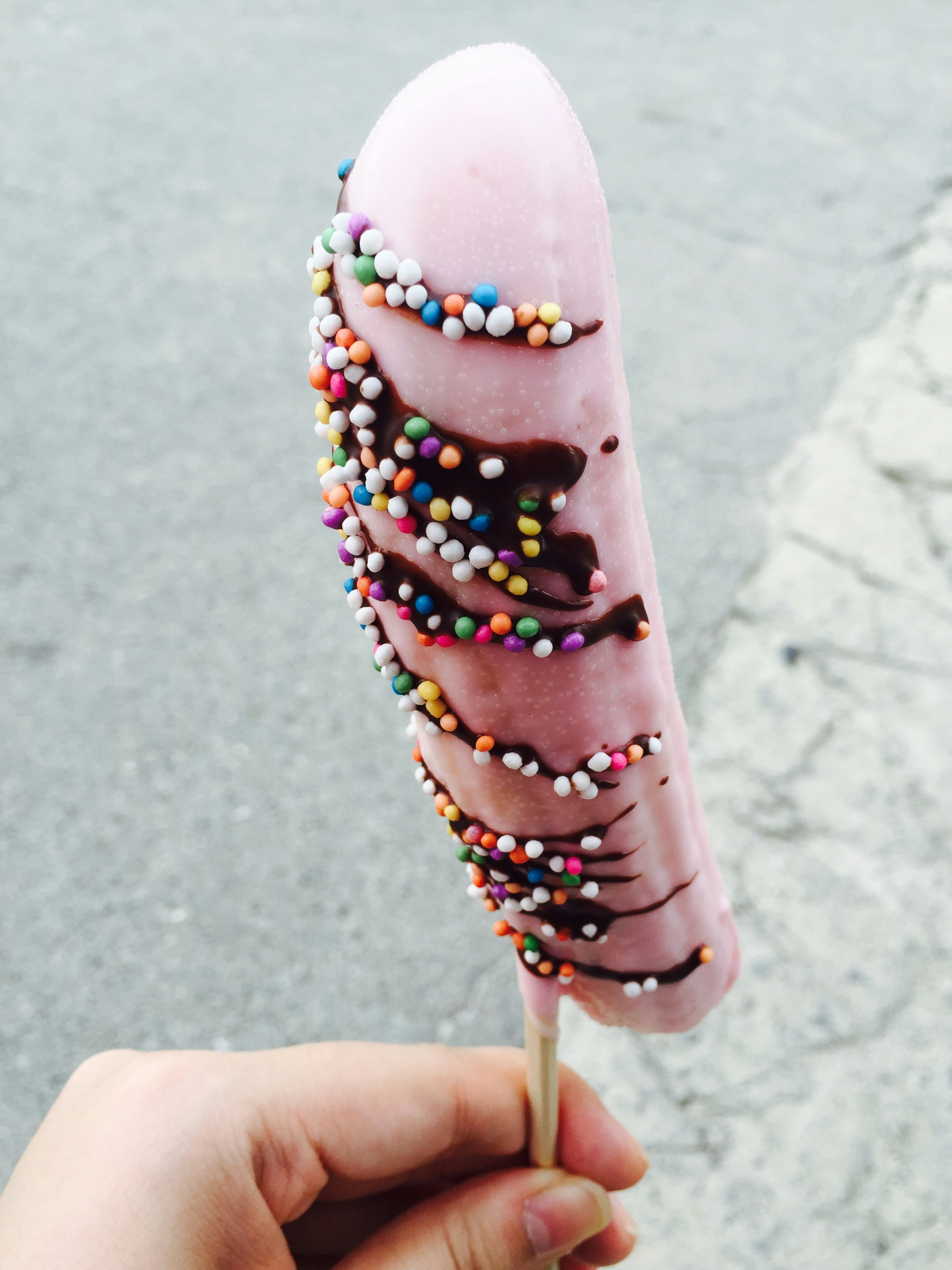 Es krim batang berwarna pink dengan taburan sprinkles warna-warni dan saus cokelat