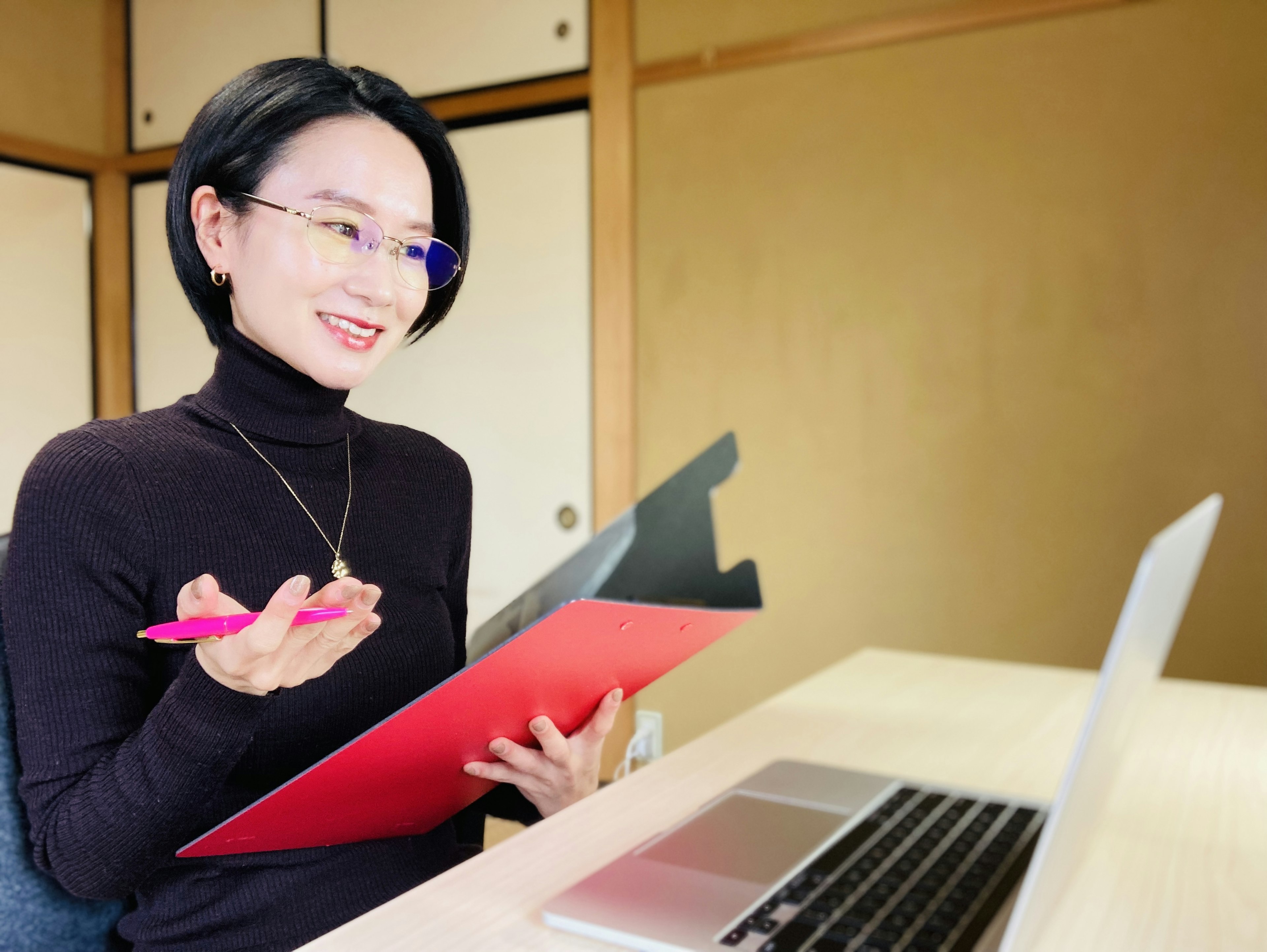 A woman in a black turtleneck holds a red clipboard smiling at a laptop