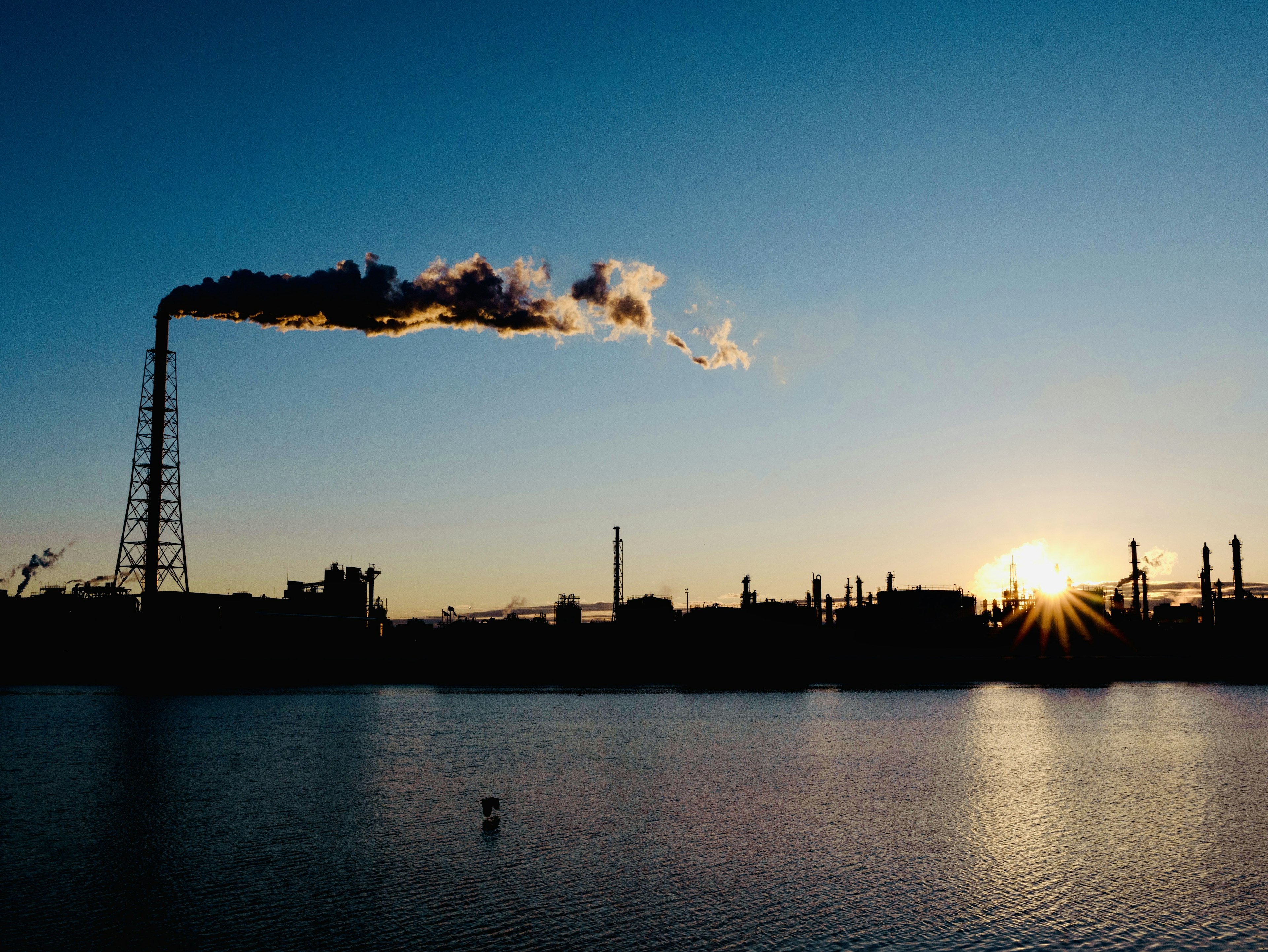 Ciminiere di fabbrica che rilasciano fumi contro un tramonto riflesso sull'acqua