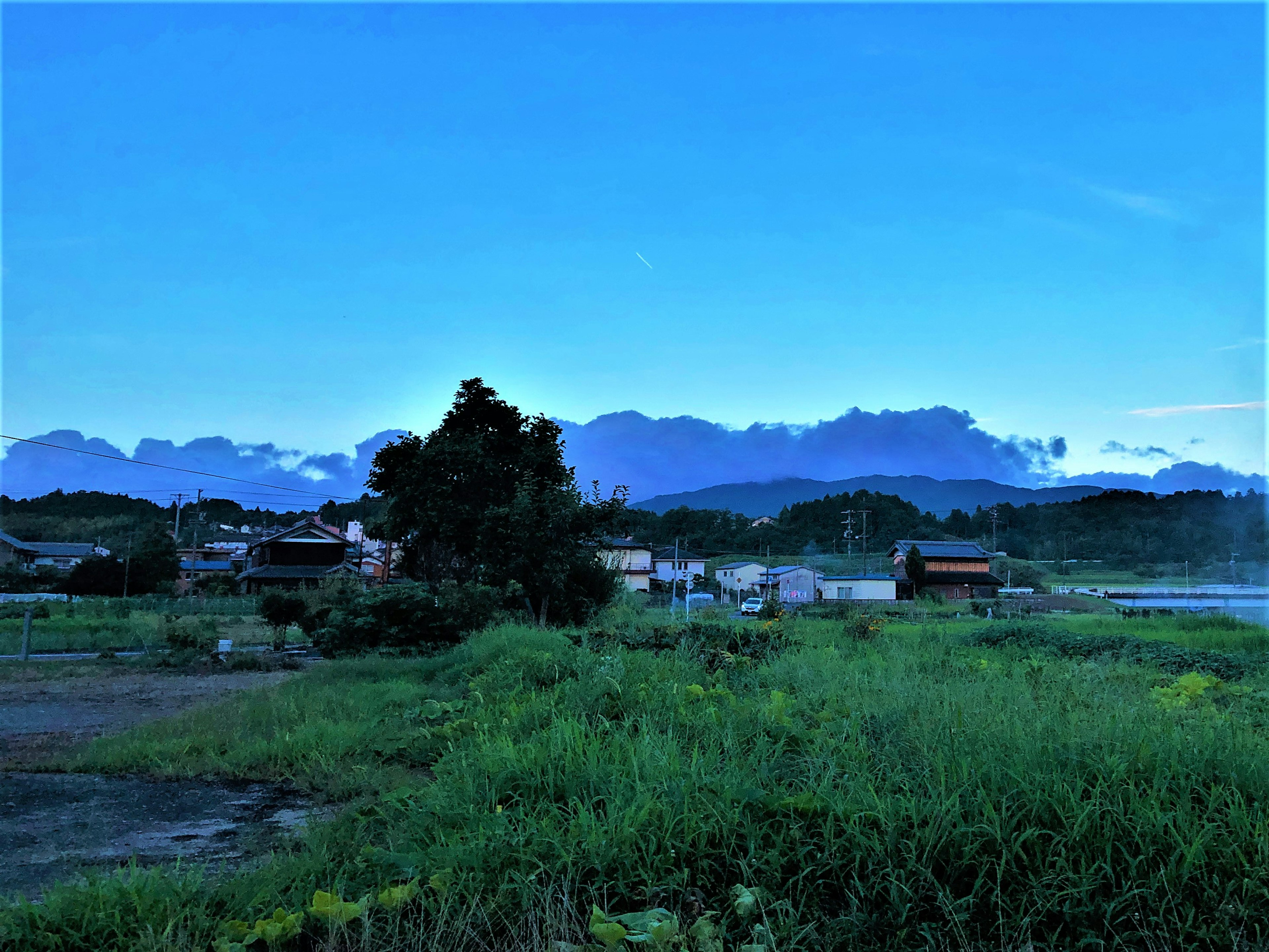 Pemandangan pedesaan dengan langit biru dan pegunungan di latar belakang menampilkan sawah dan rumah kecil