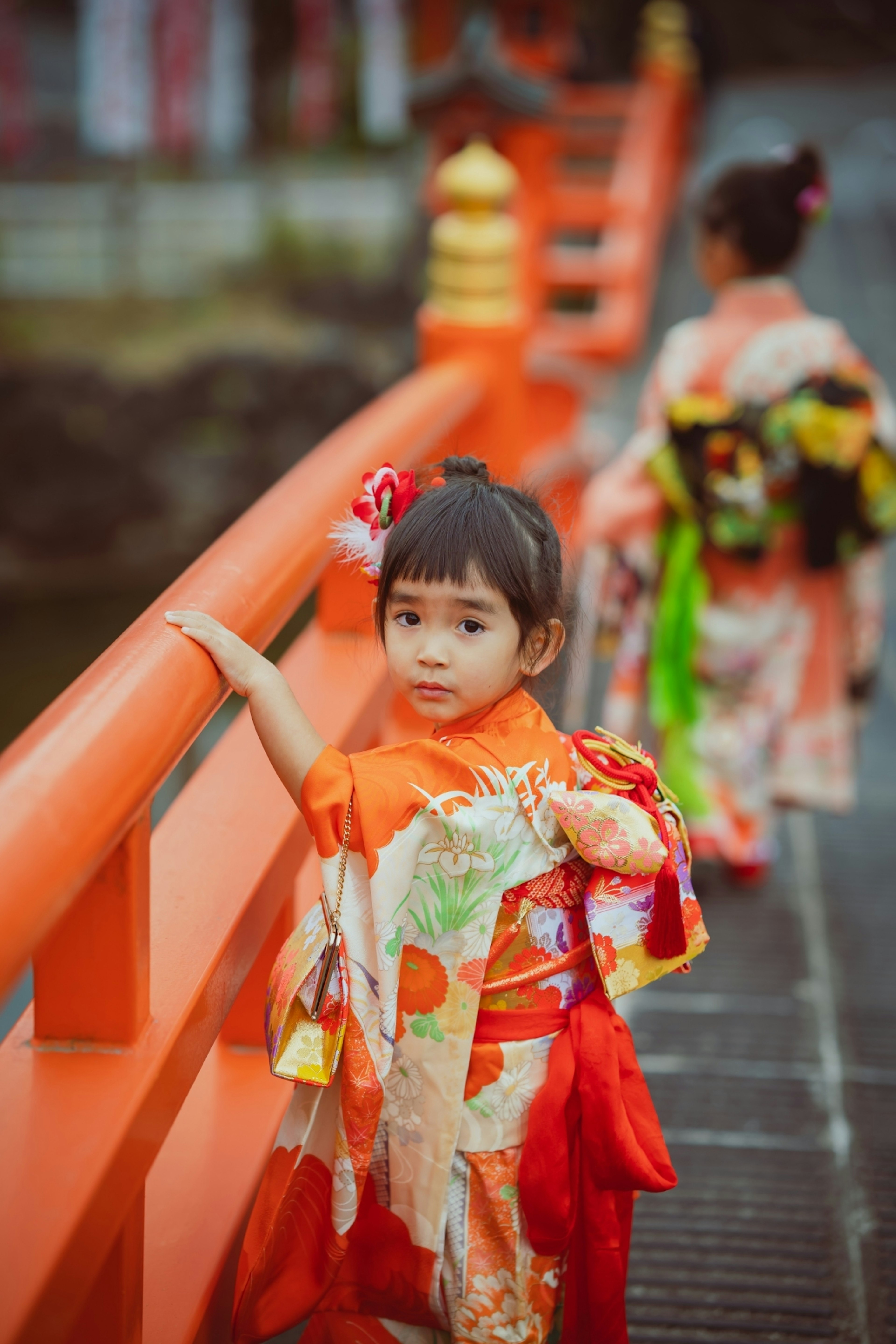 Ein Kind in einem lebhaften Kimono steht auf einer orangefarbenen Brücke mit einem anderen Kind im Hintergrund