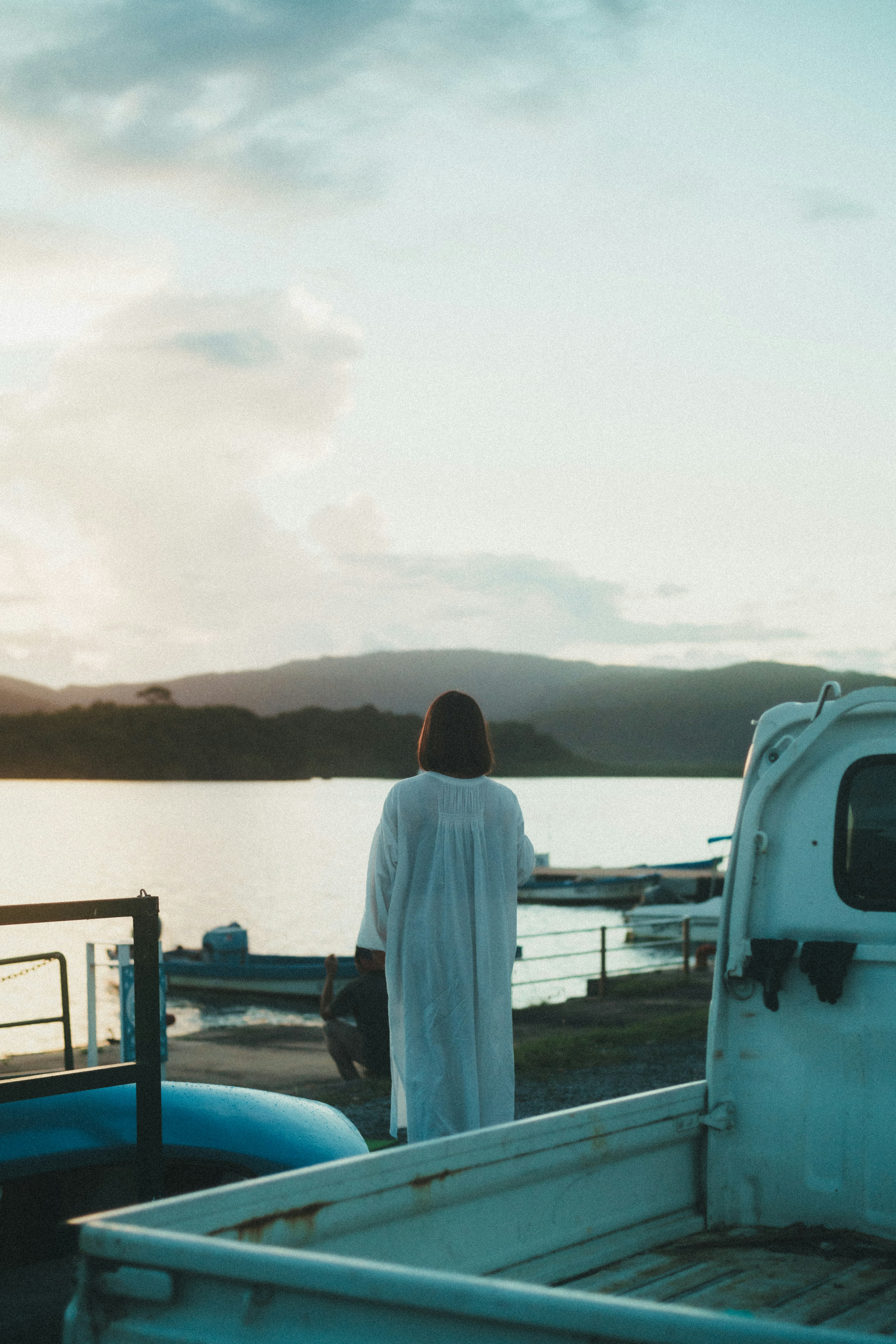 Una mujer con un vestido blanco está de espaldas junto a un lago sereno al atardecer