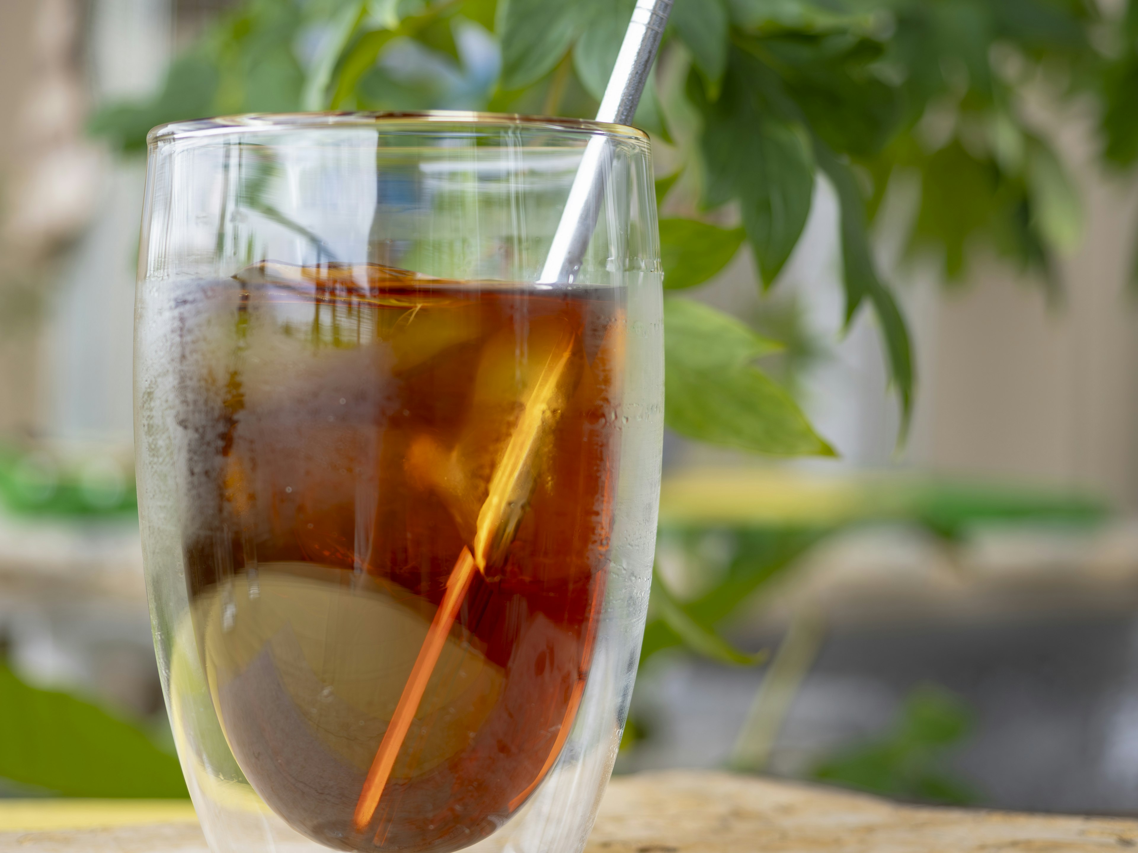 A glass of iced tea with a straw and greenery in the background