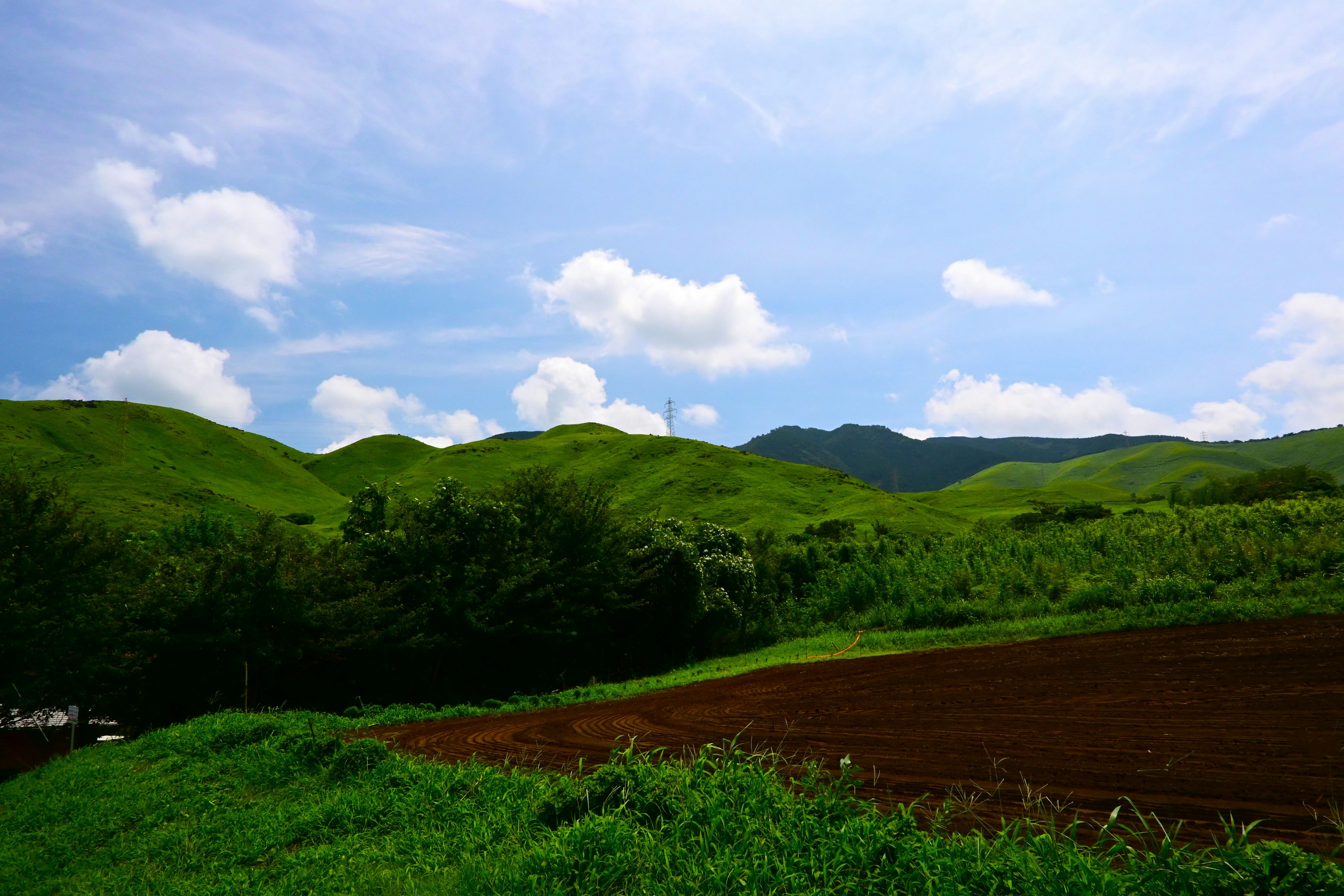 Üppige grüne Hügel unter einem klaren blauen Himmel mit verstreuten Wolken und sichtbarem Ackerland