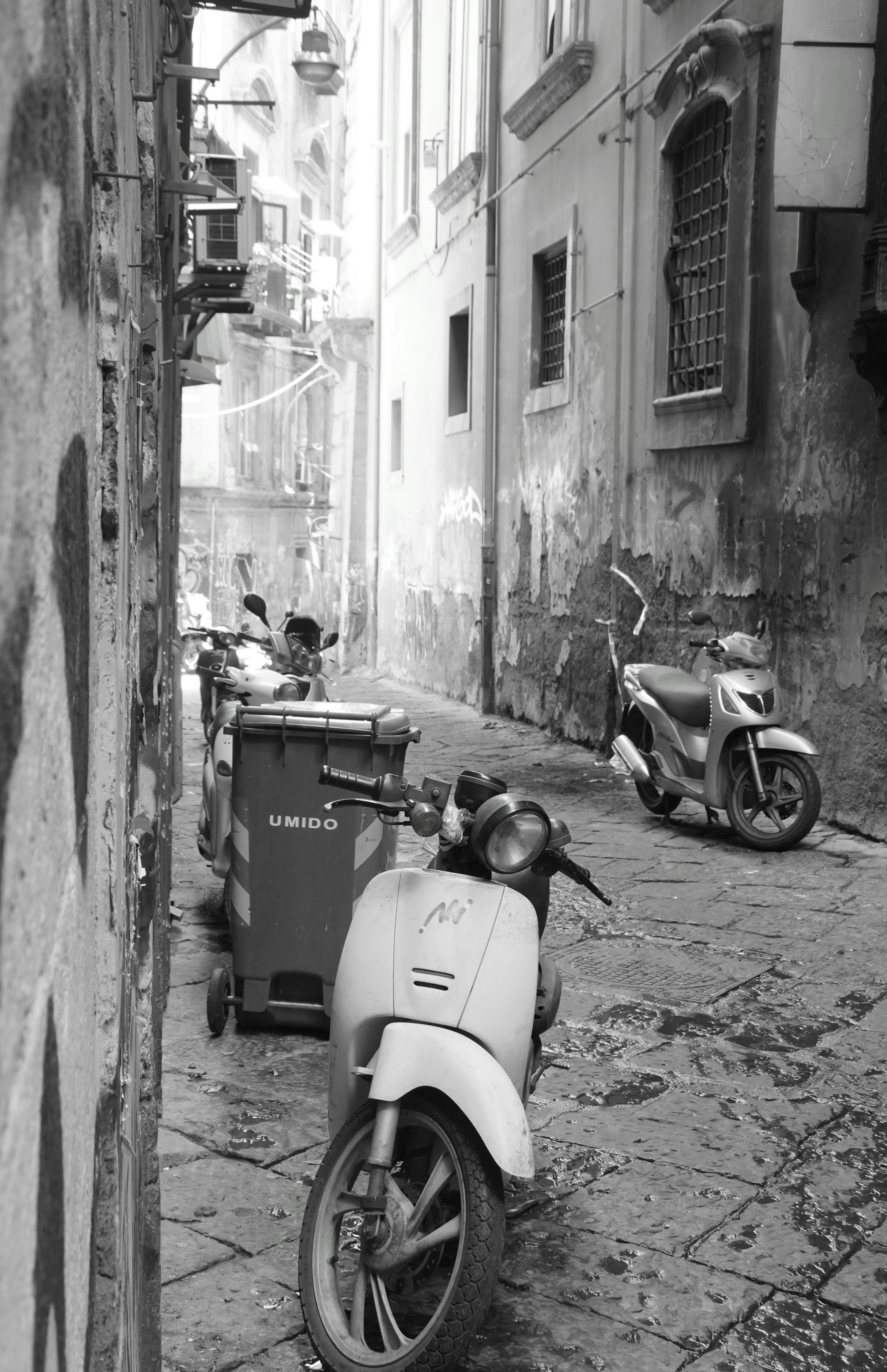 Black and white photo of scooters and a trash bin in a narrow street