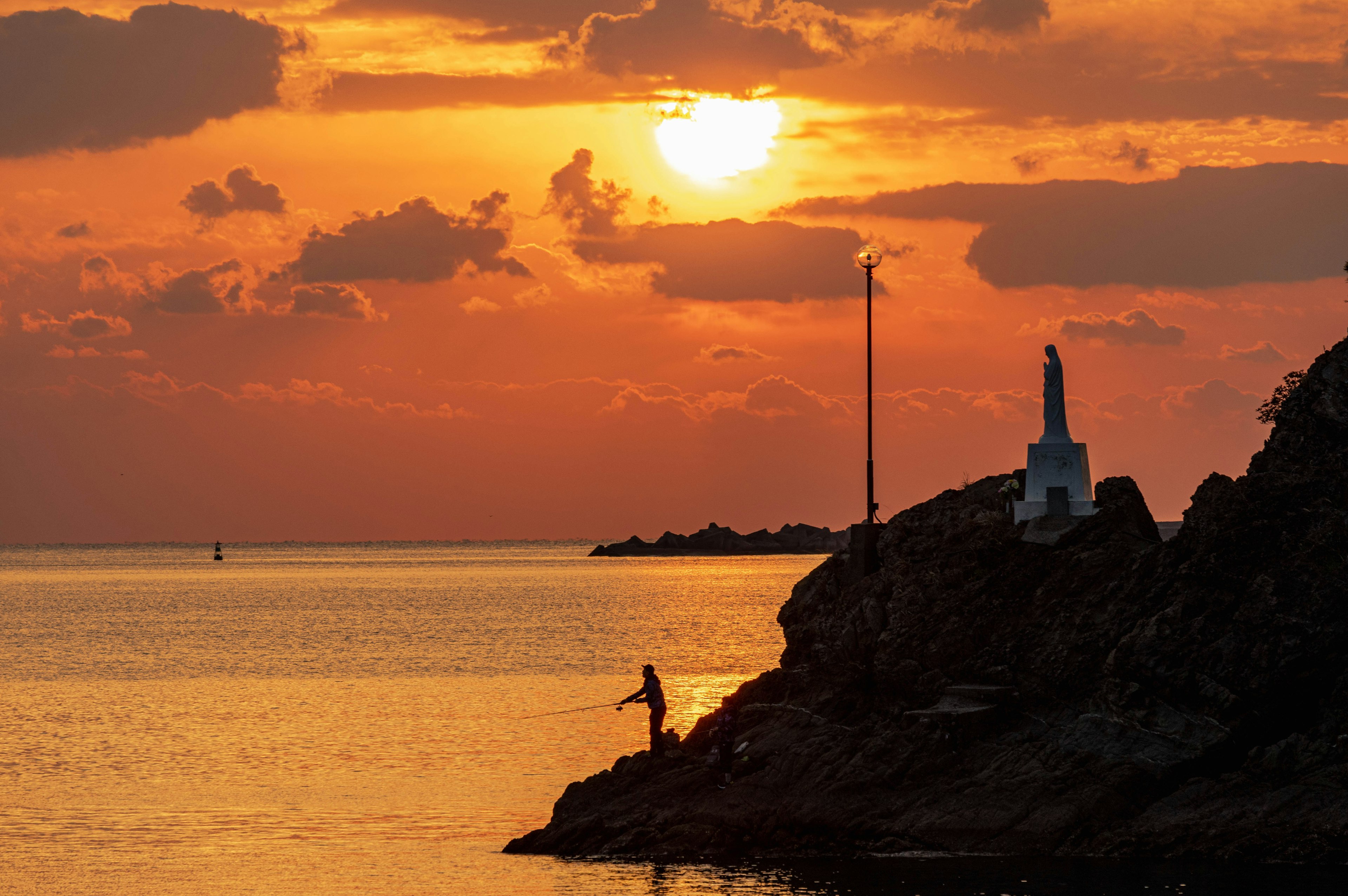 Fisherman standing on rocky shore with sunset and statue in background