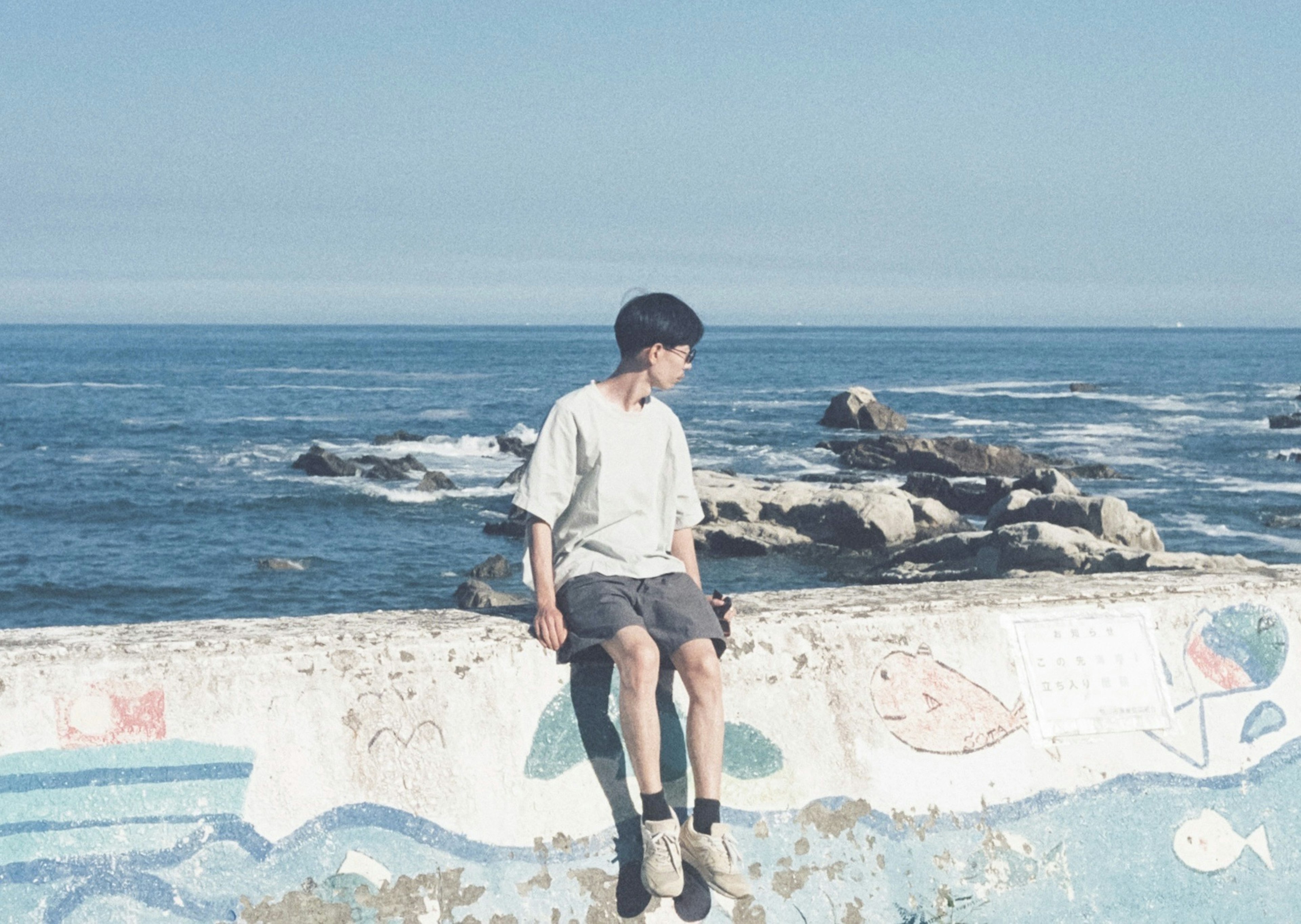 Ein Junge sitzt auf einer Mauer am Meer mit blauem Ozean und Himmel im Hintergrund