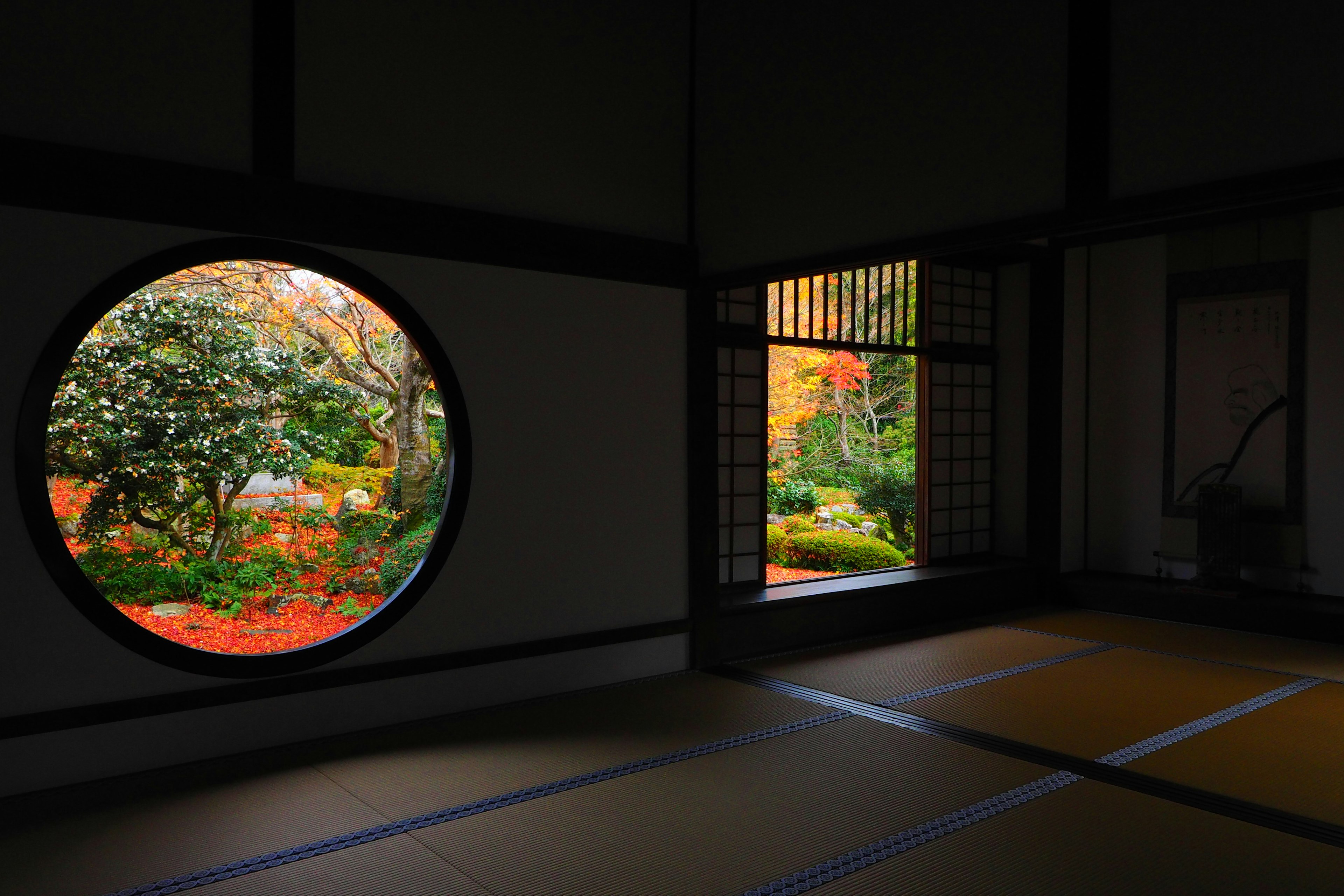 Innenraum eines traditionellen japanischen Zimmers mit einem runden Fenster, das Herbstlaub zeigt, und einem quadratischen Fenster mit Blick auf den Garten