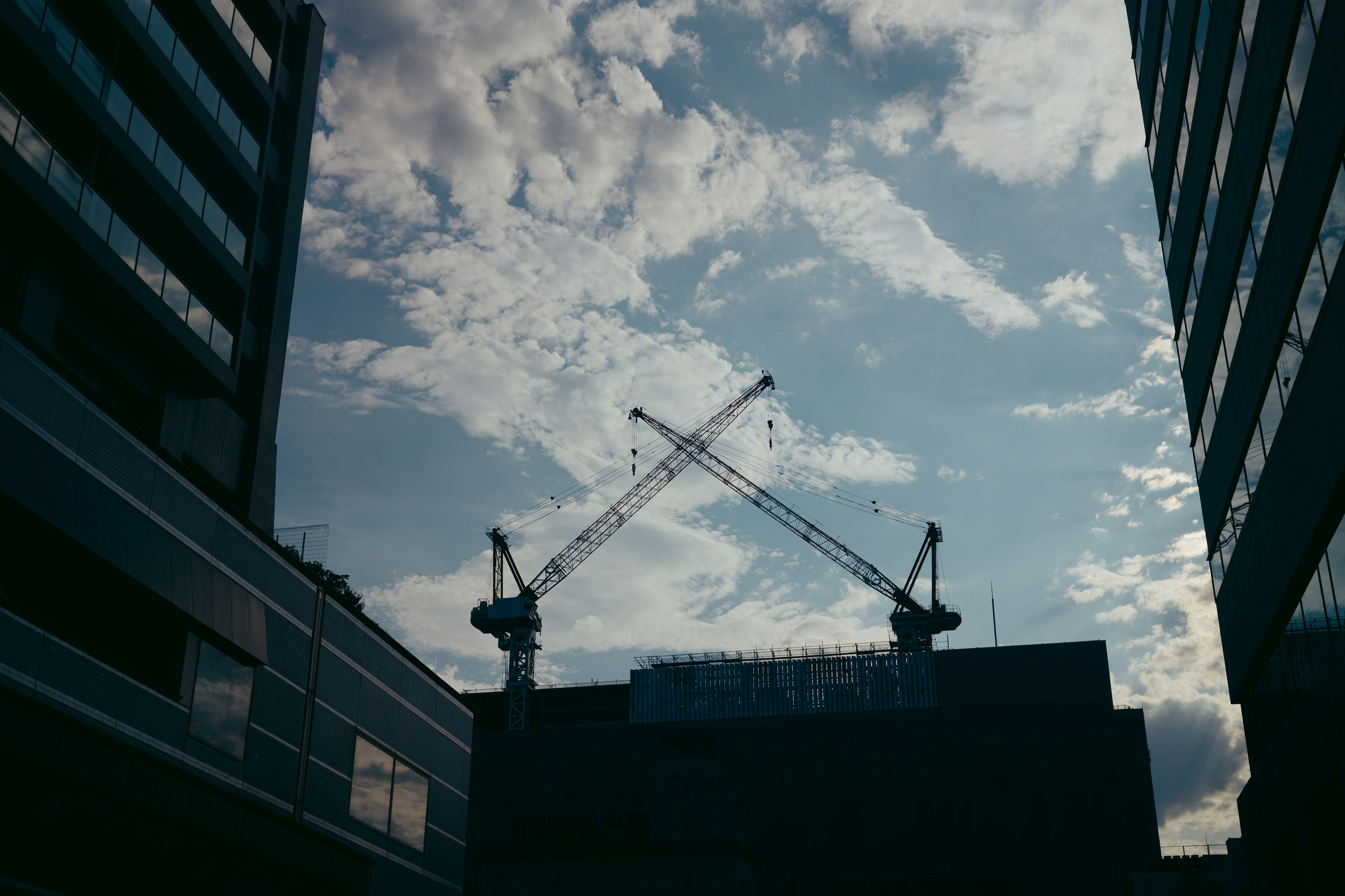 Silhouette of intersecting cranes against a blue sky with clouds between buildings