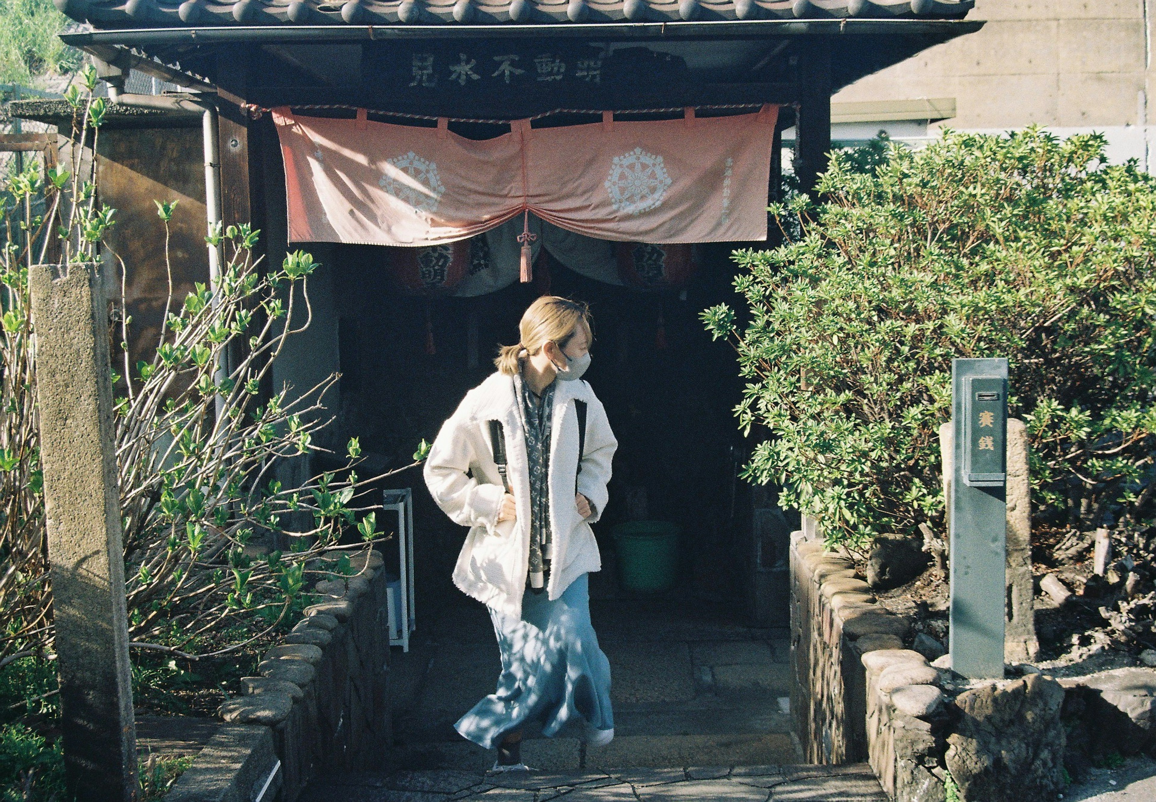 A woman walking at the entrance of a traditional building surrounded by greenery and a warm-colored curtain