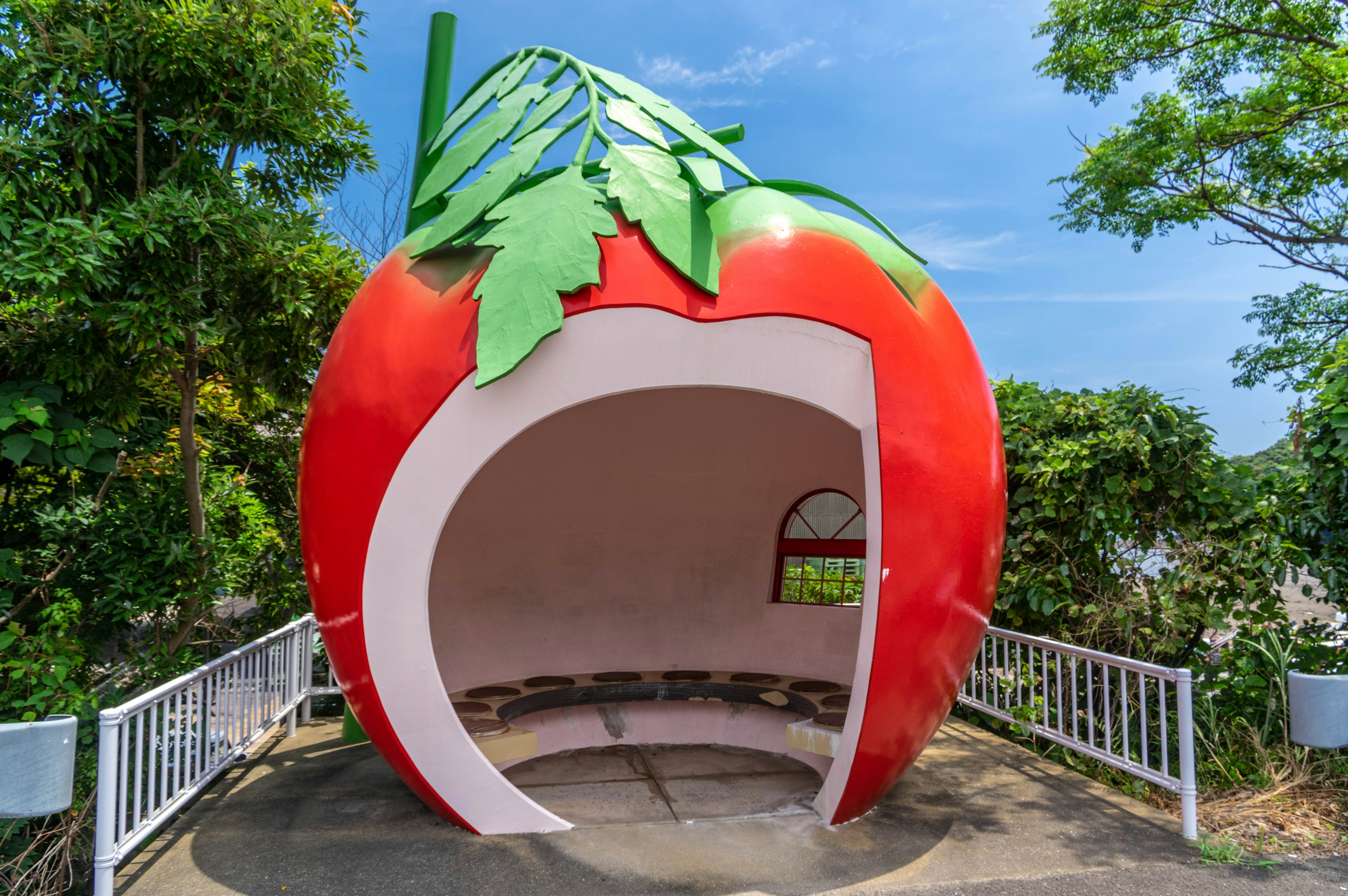 Ein großes rotes Erdbeer-förmiges Schutzdach mit grünen Blättern umgeben von Bäumen und blauem Himmel