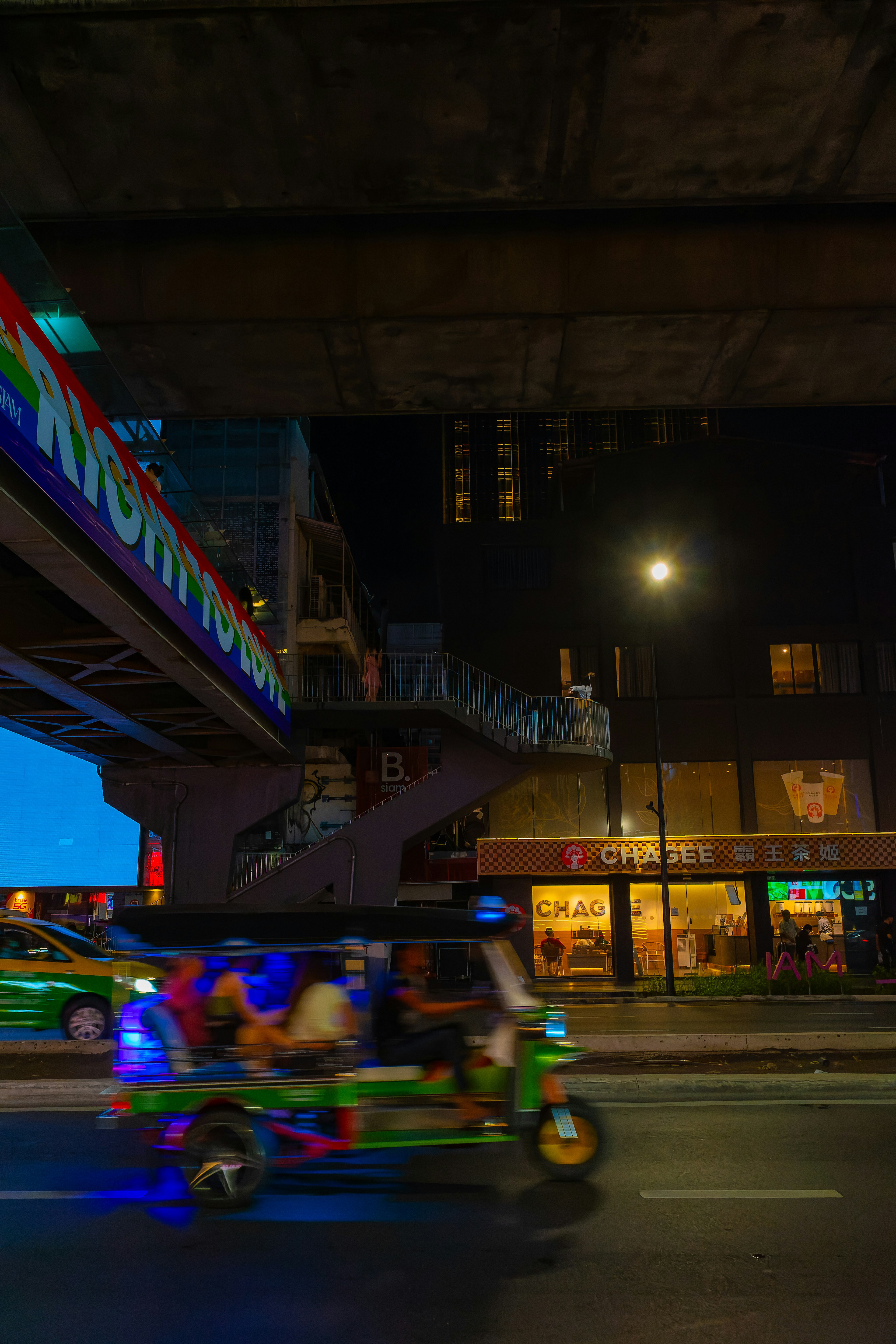 A tuk-tuk driving through a vibrant street at night with colorful signage and shop lights under a bridge