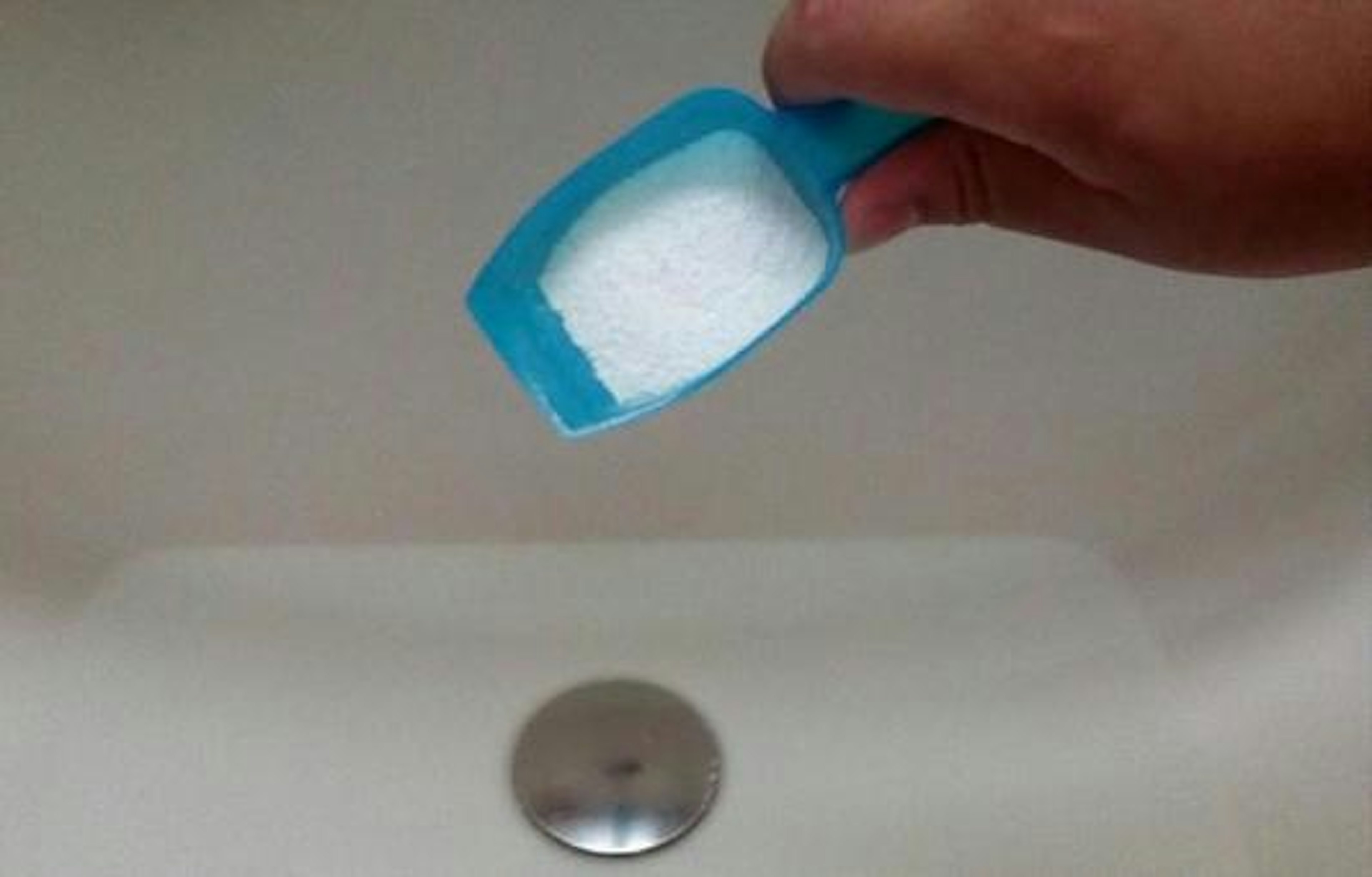 A hand holding a blue scoop filled with white powder above a sink drain